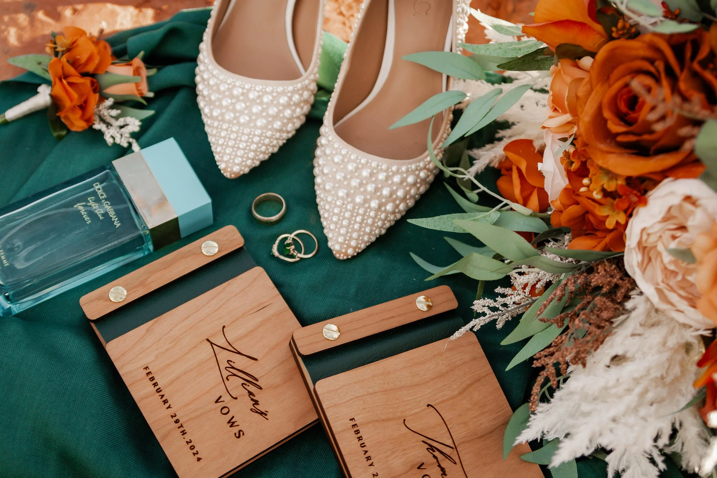 Wedding table setting with pearl embellished high heel shoes, a bouquet of orange and white flowers, a bottle of Dolce & Gabbana perfume, a gold wedding band, two wedding rings, and small wooden signs with names and wedding date on a dark green cloth