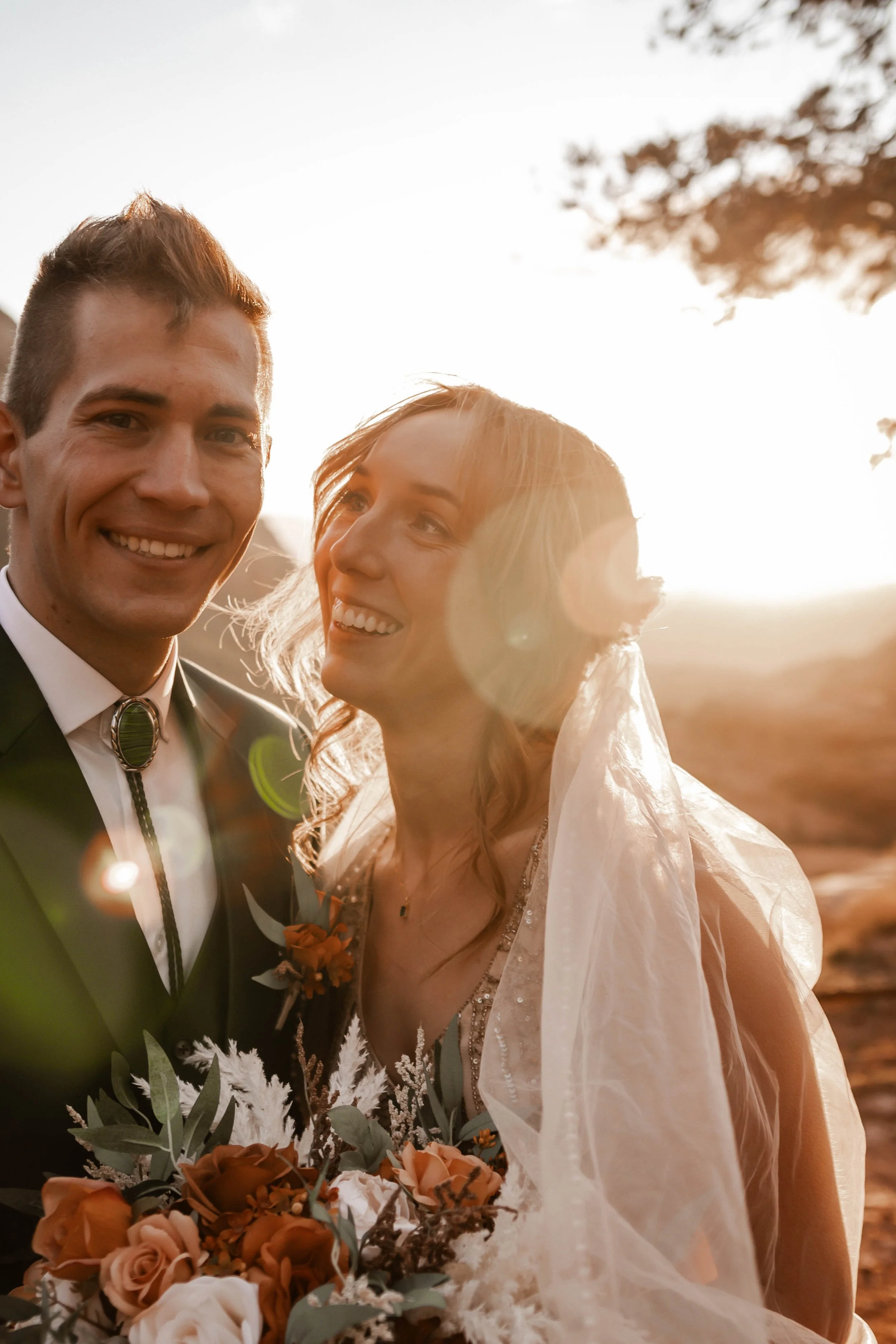 A happy couple on their wedding day, standing outdoors during sunset with the bride wearing a veil and holding a bouquet of flowers.
