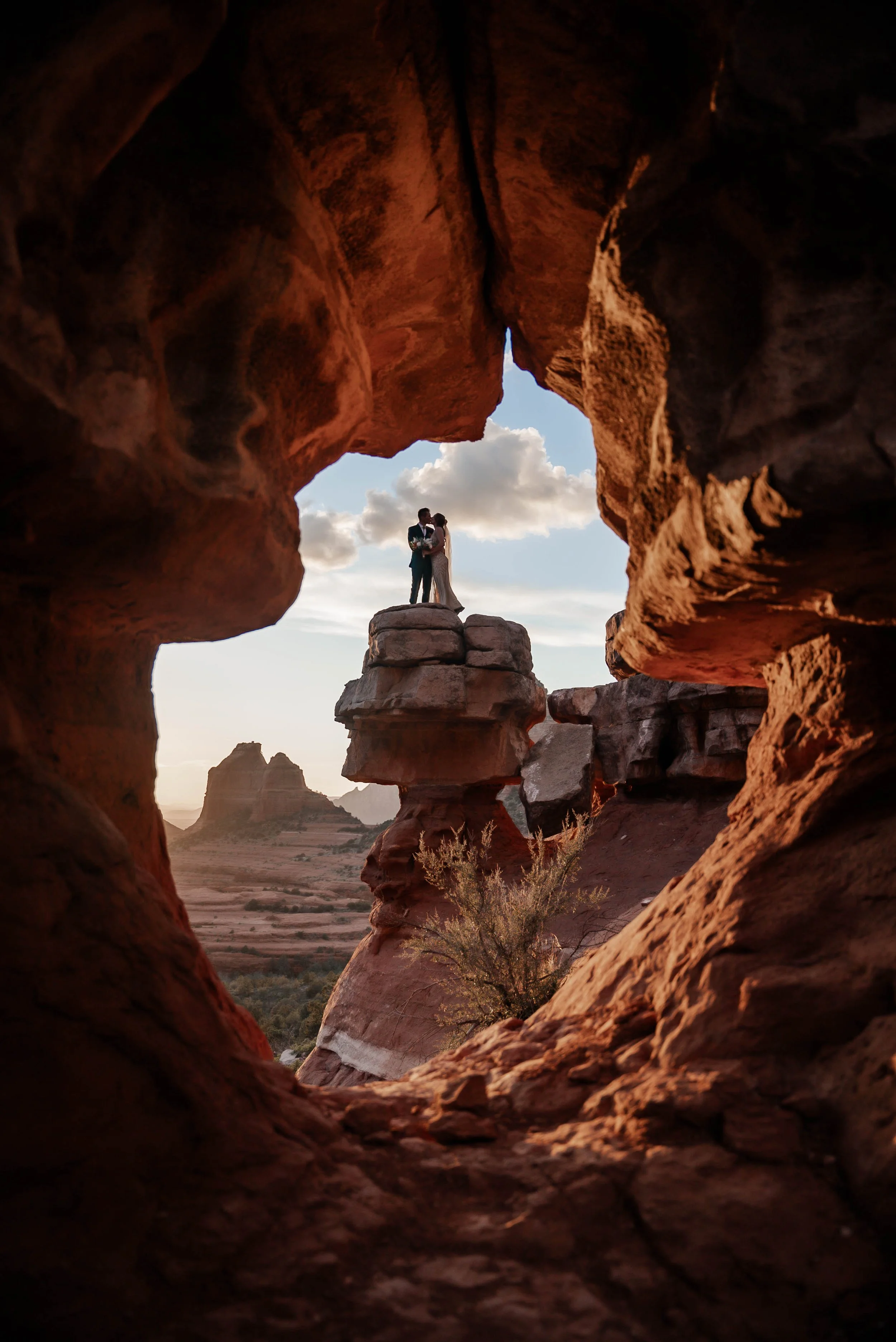 A silhouette of a couple standing on a large rock formation in a desert landscape, framed by a rock window, with a cloudy sky and distant mesas in the background.