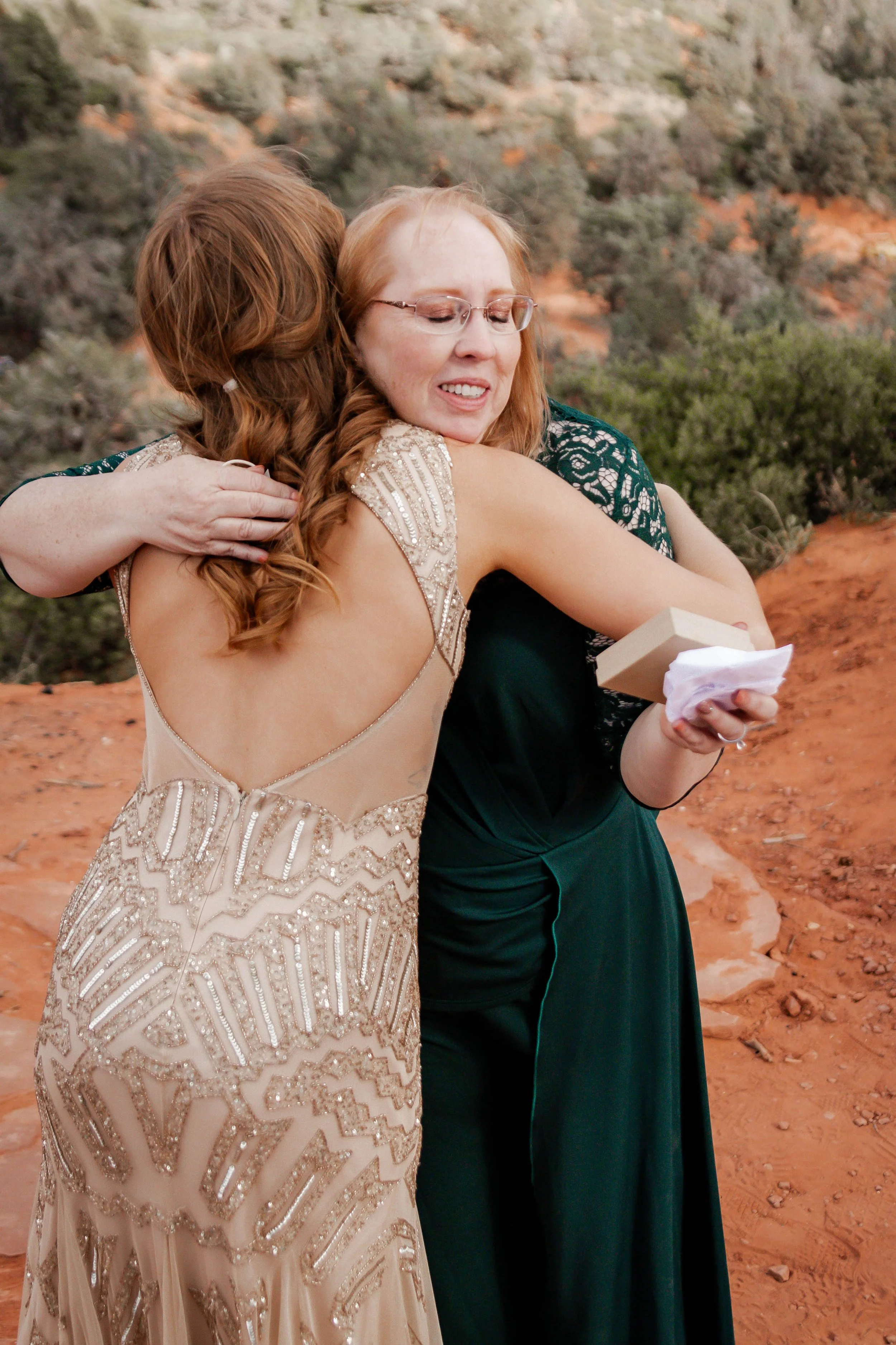 Two women hugging, one in a beige, beaded dress and the other in a dark green dress, outdoors with desert landscape in the background.