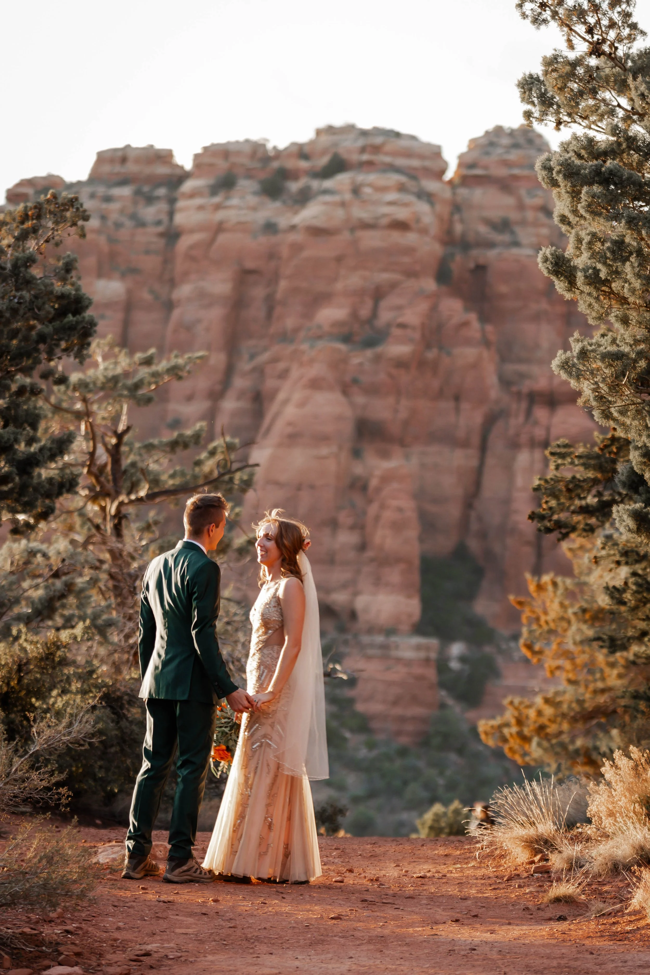 A couple in wedding attire holding hands in a desert landscape with large red rock formations and trees in Sedona, Arizona.