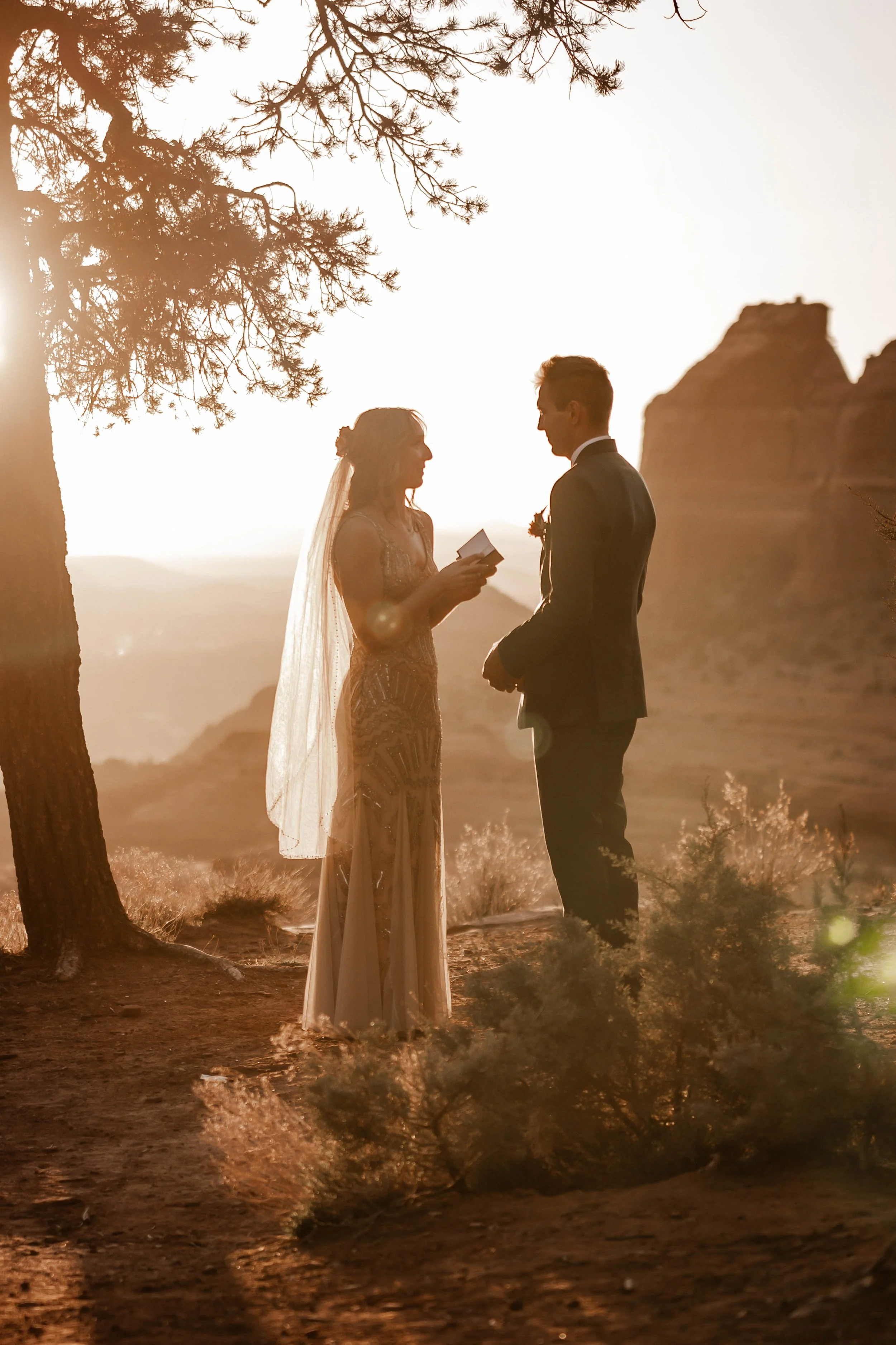 A bride and groom exchanging vows outdoors at sunset in a desert landscape, with trees and rock formations in the background.