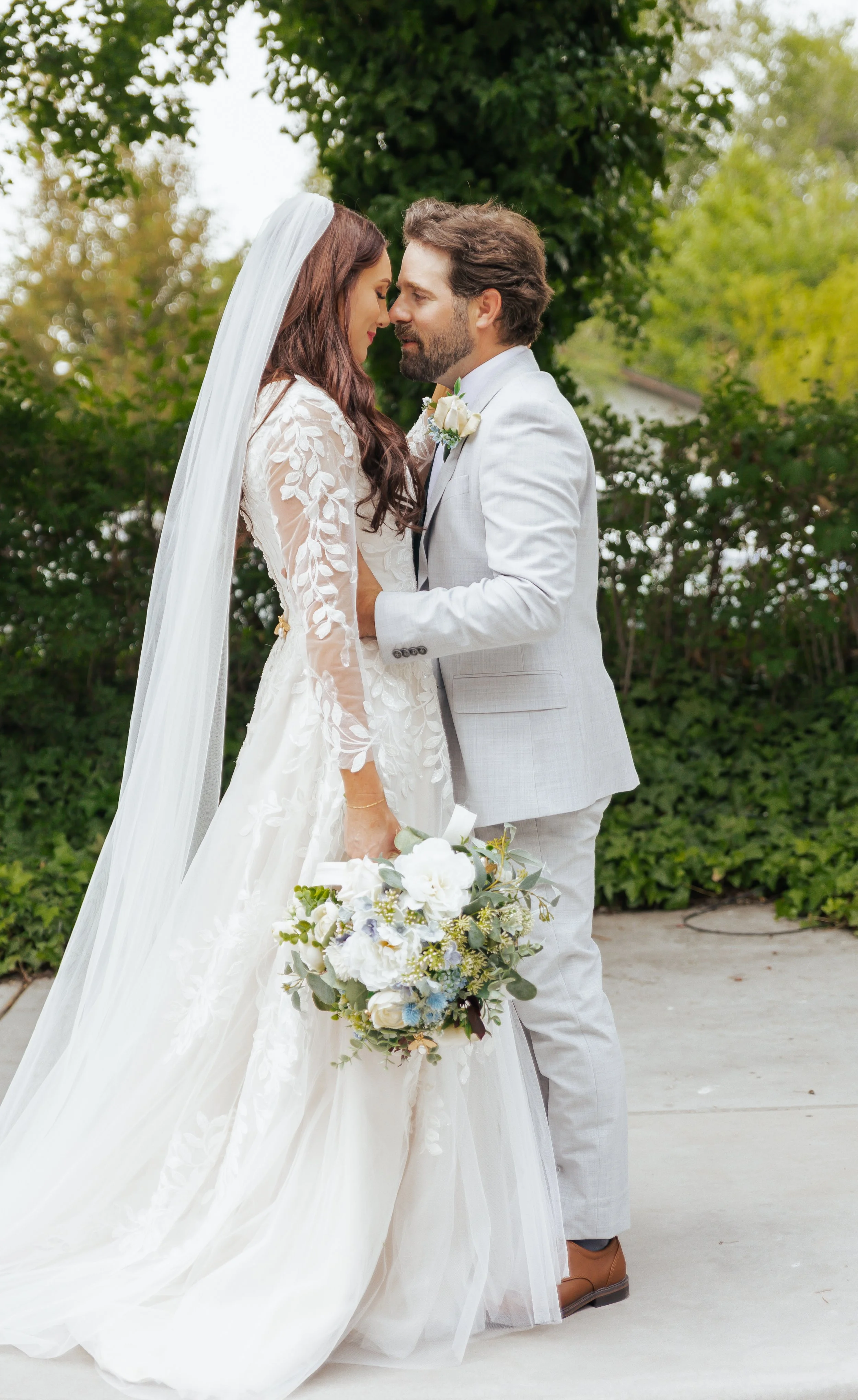 A bride and groom stand close together outdoors, foreheads touching, holding a bouquet of flowers, surrounded by green trees.