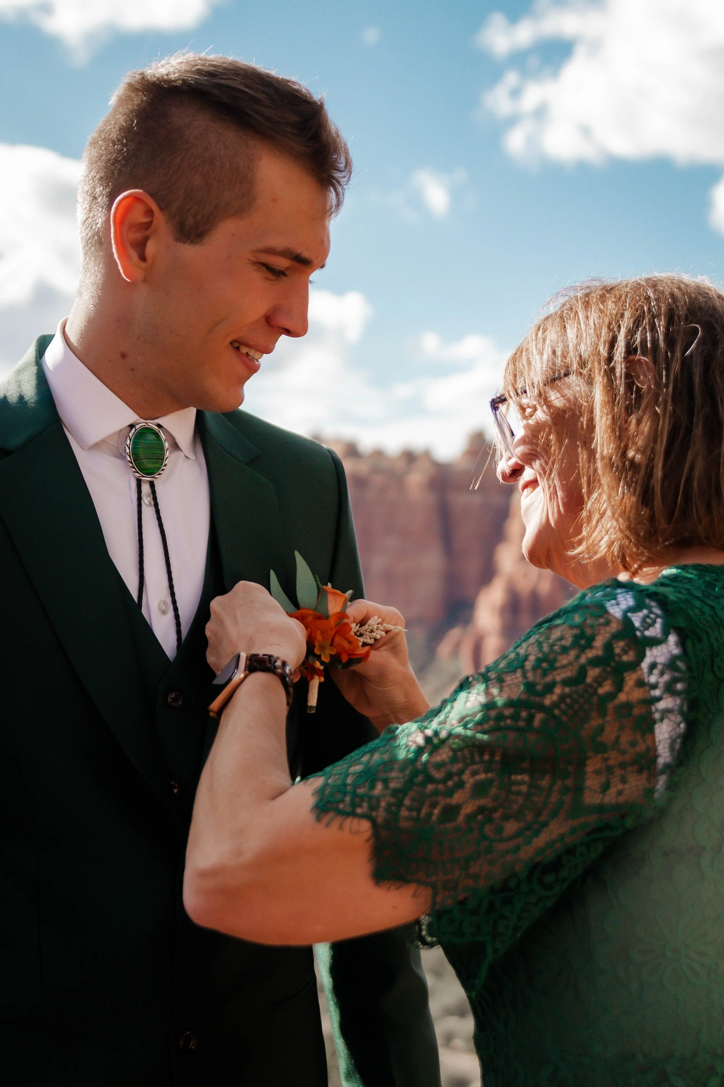 A young man in a green suit is being pinned with a corsage by an older woman in a green lace dress outdoors against a background of red rock cliffs and blue sky.
