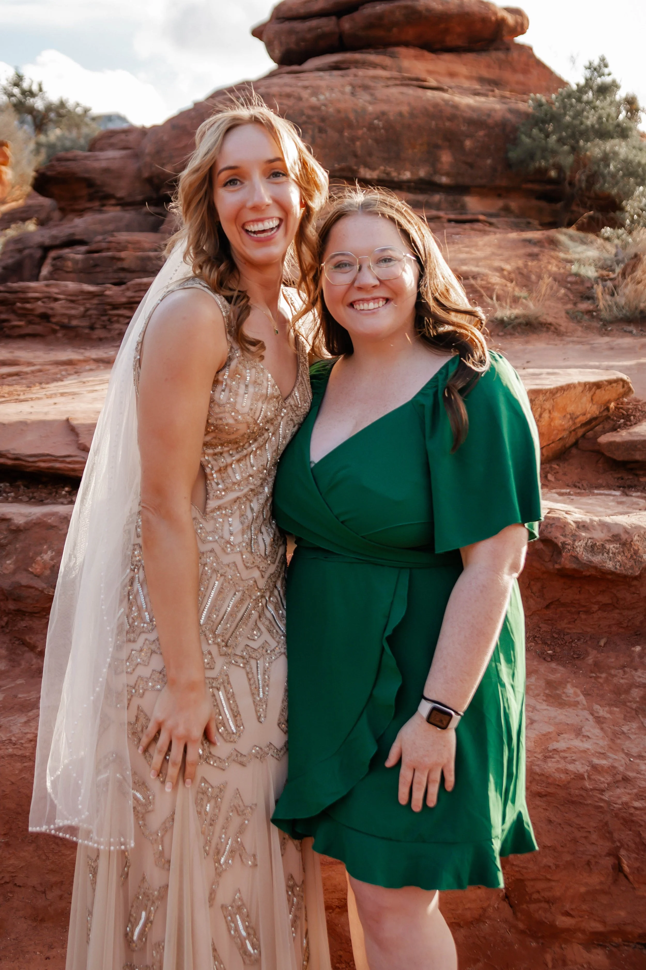 Two women in formal dresses smiling and posing outdoors with red rock formations in the background.