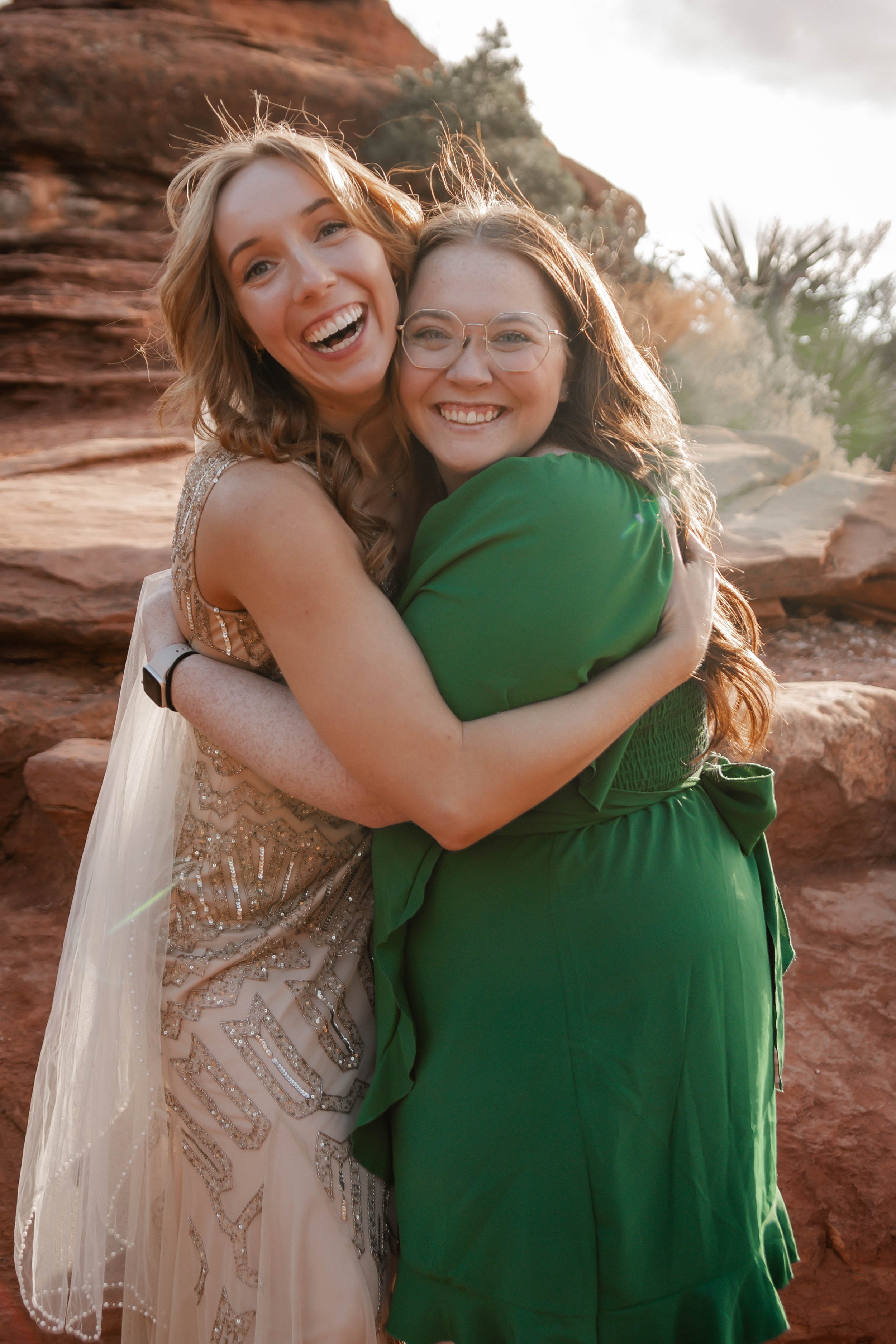 Two women smiling and hugging outdoors with desert rocks and plants in the background.