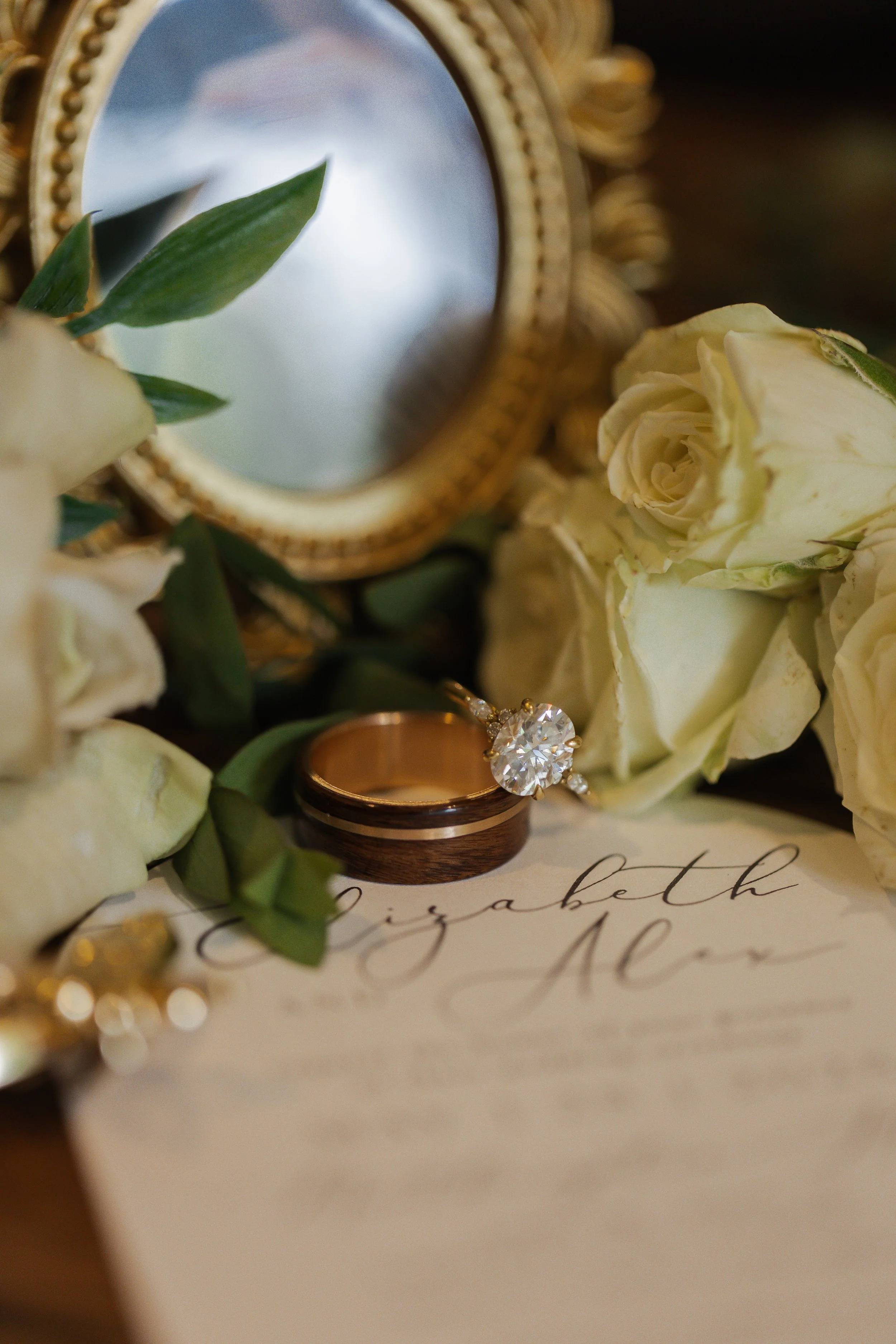 A gold wedding ring, a diamond engagement ring, white roses, and green leaves placed near a mirror and a handwritten invitation.