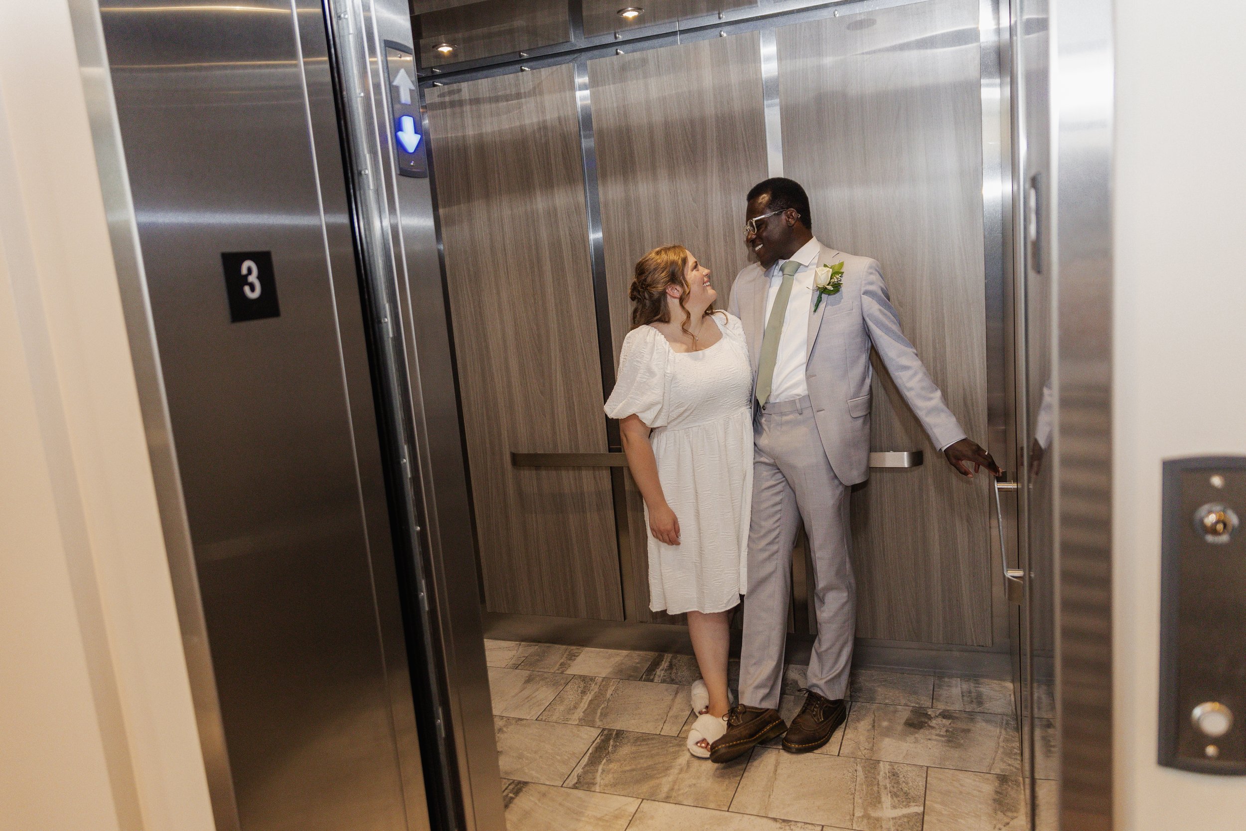 A couple in wedding attire sharing a moment in an elevator, with the bride in a white dress and the groom in a light suit, smiling at each other.