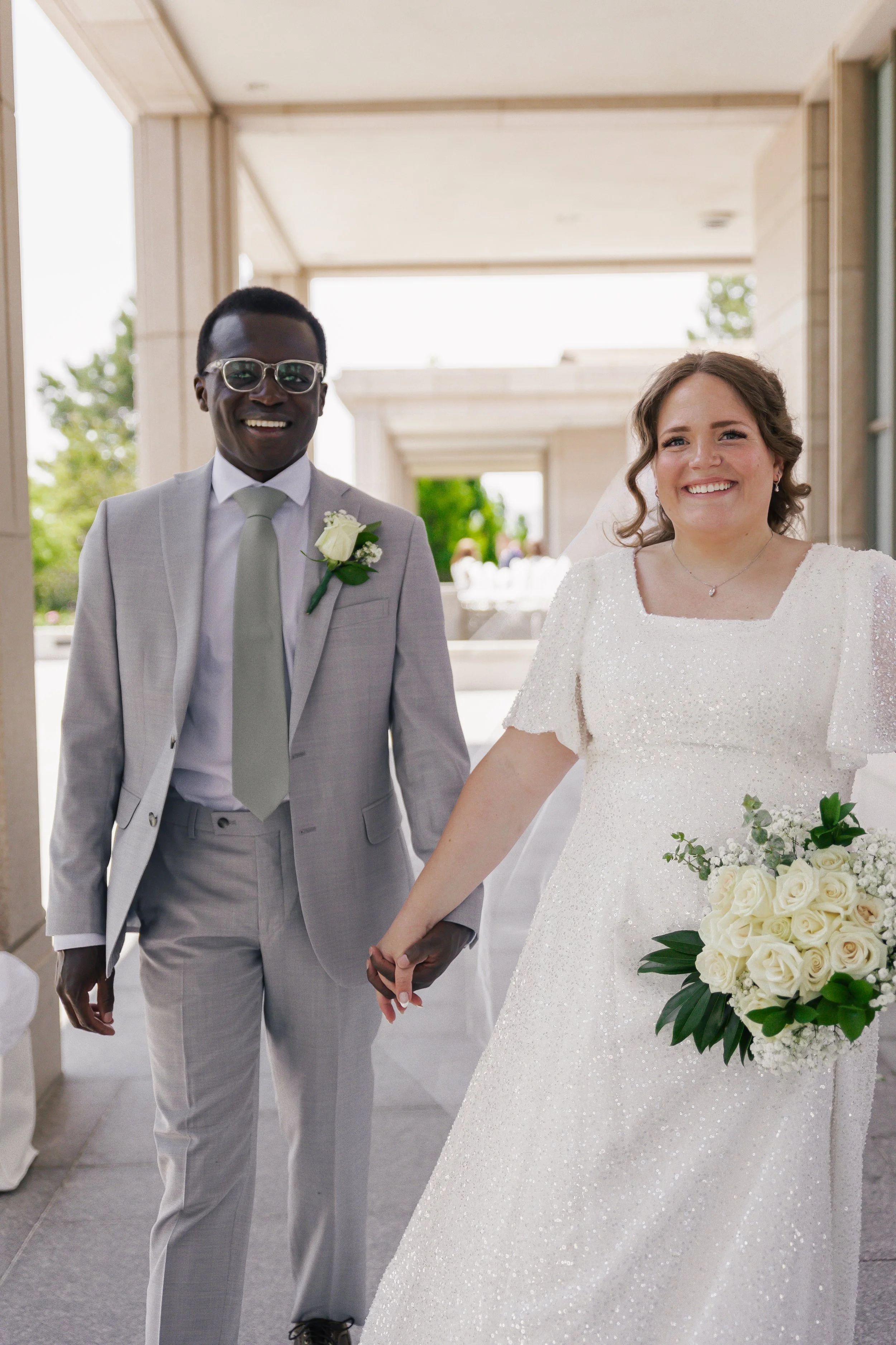 A newly married couple holding hands and smiling outdoors, with the groom in a light gray suit and the bride in a white wedding dress holding a bouquet of white roses.