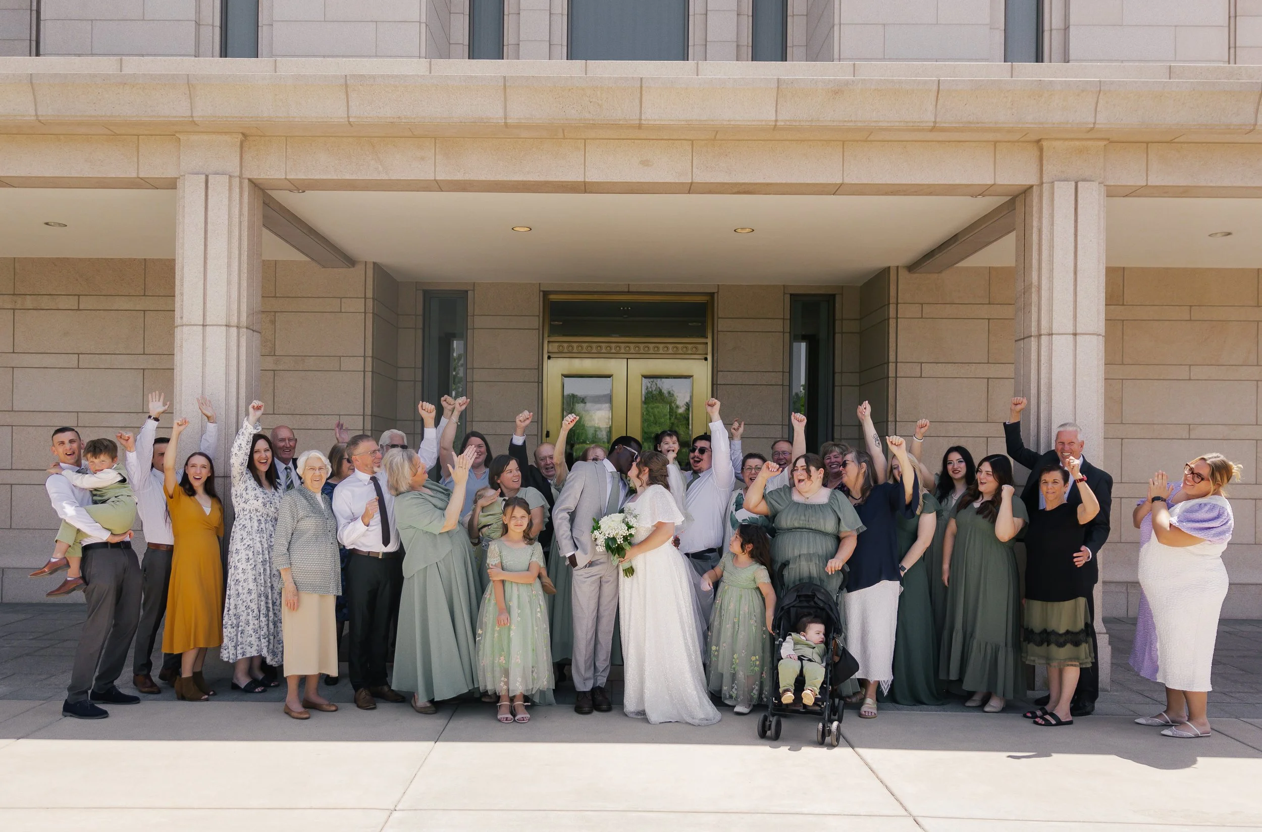 Group of people celebrating outside a building, including a bride and groom, family members, and children, all raising their hands in joy.