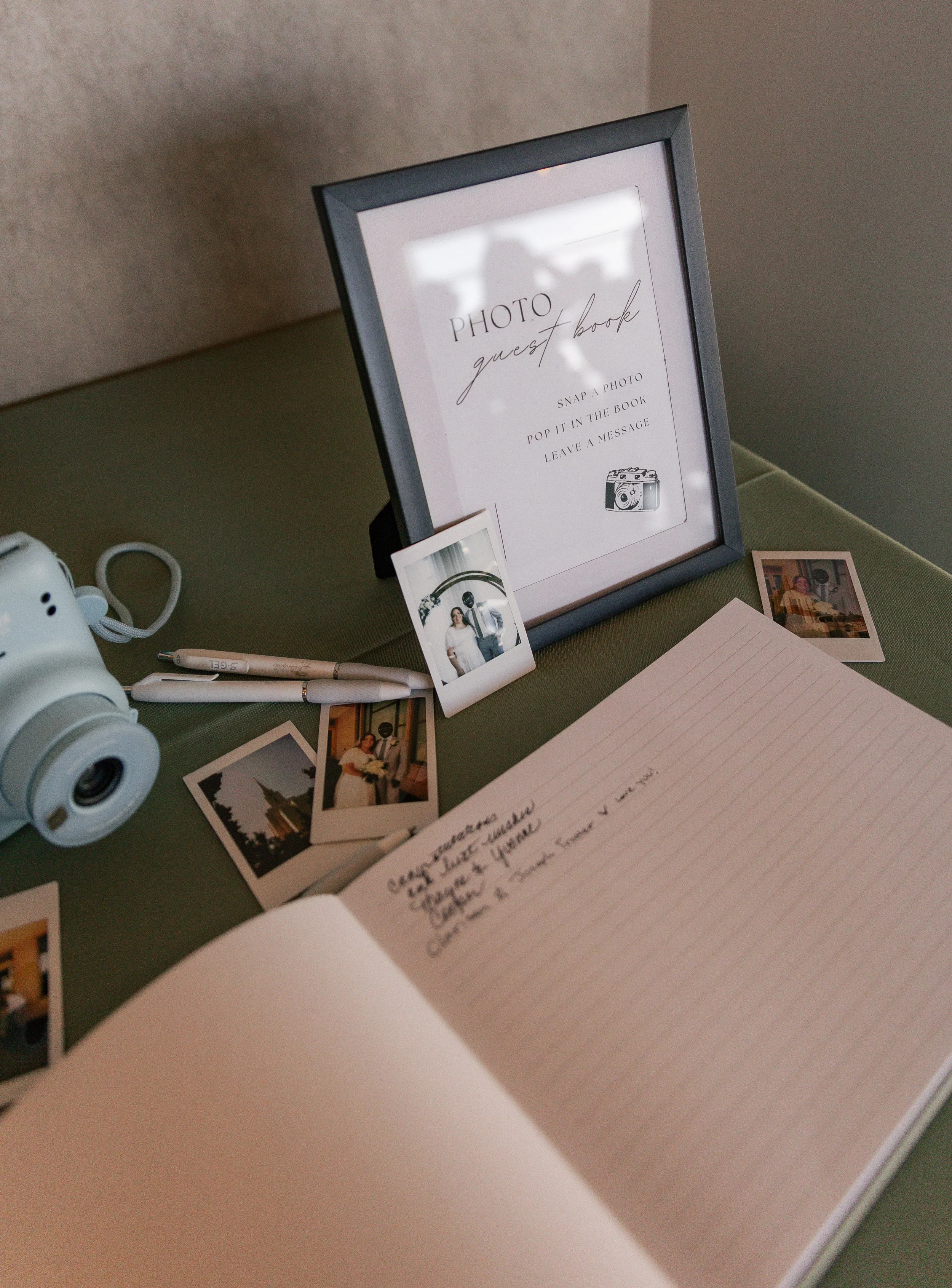 A table with a guest book, small photographs, a camera, and pens, with a sign instructing guests to snap photos, pop them in the guest book, and leave a message.