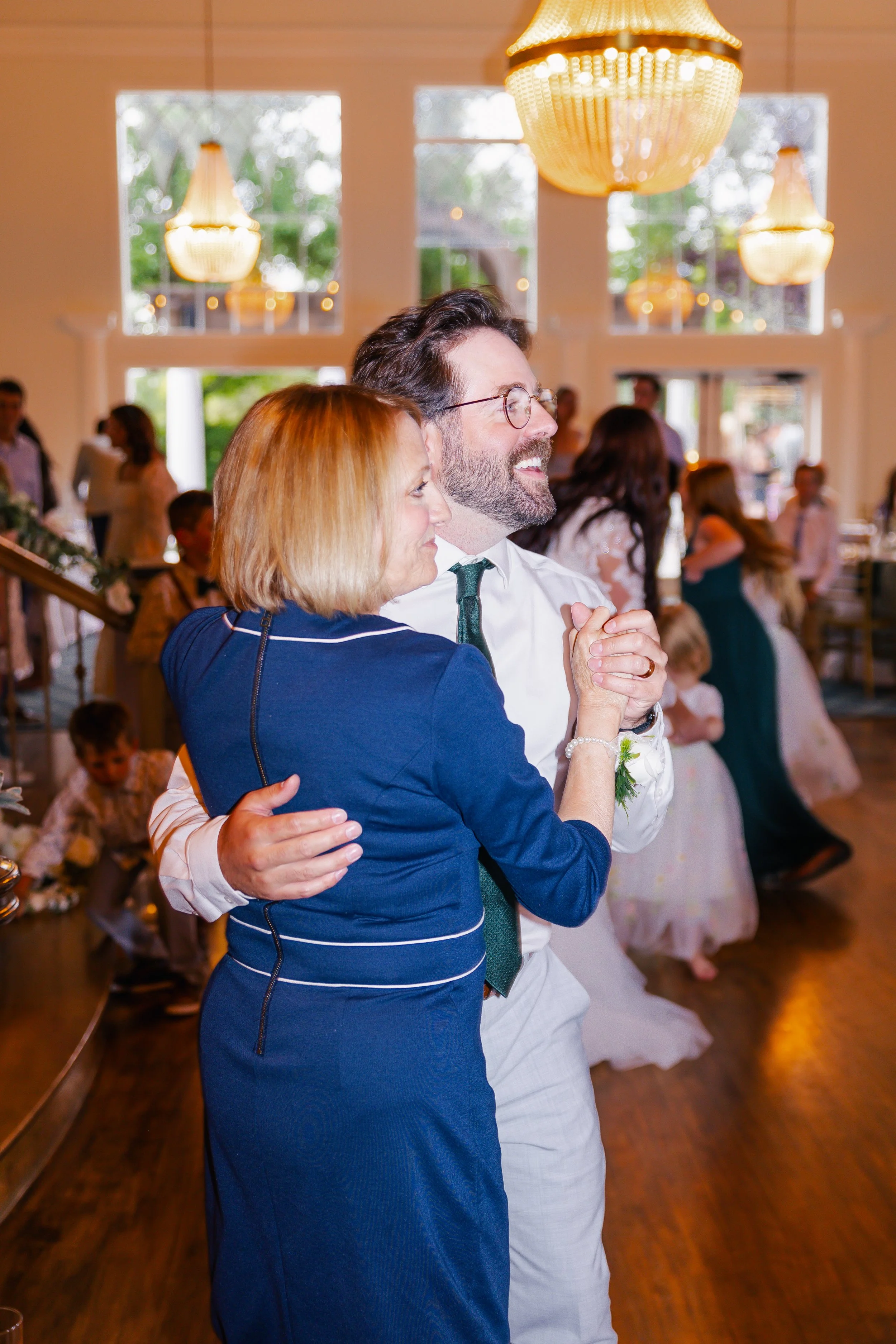 A joyful couple dancing together at a celebration, with others dancing in the background inside a well-lit room with large windows and chandeliers.