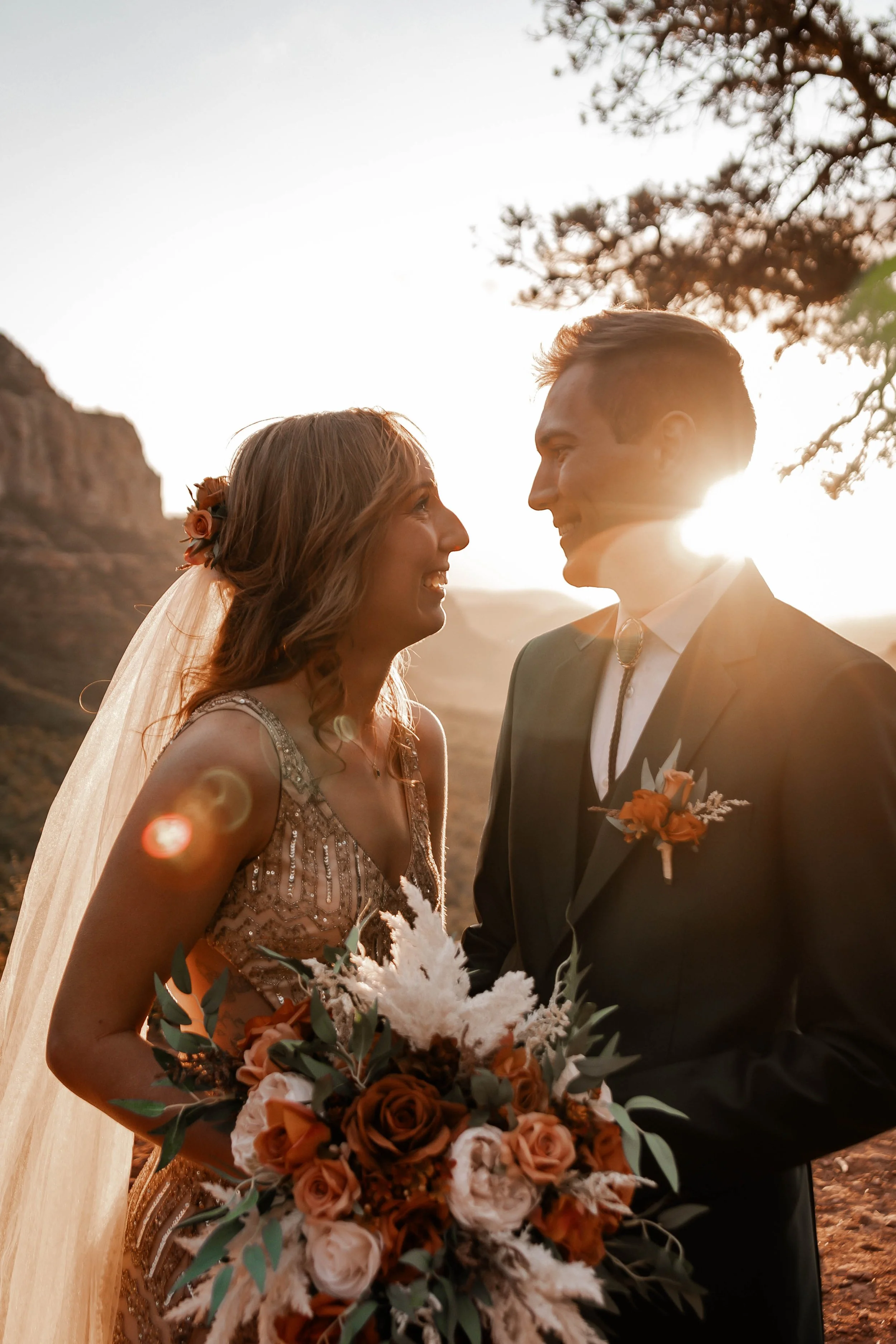 A bride and groom face each other at sunset outdoors, with the bride holding a bouquet of flowers, and the groom wearing a dark suit with an orange boutonniere.