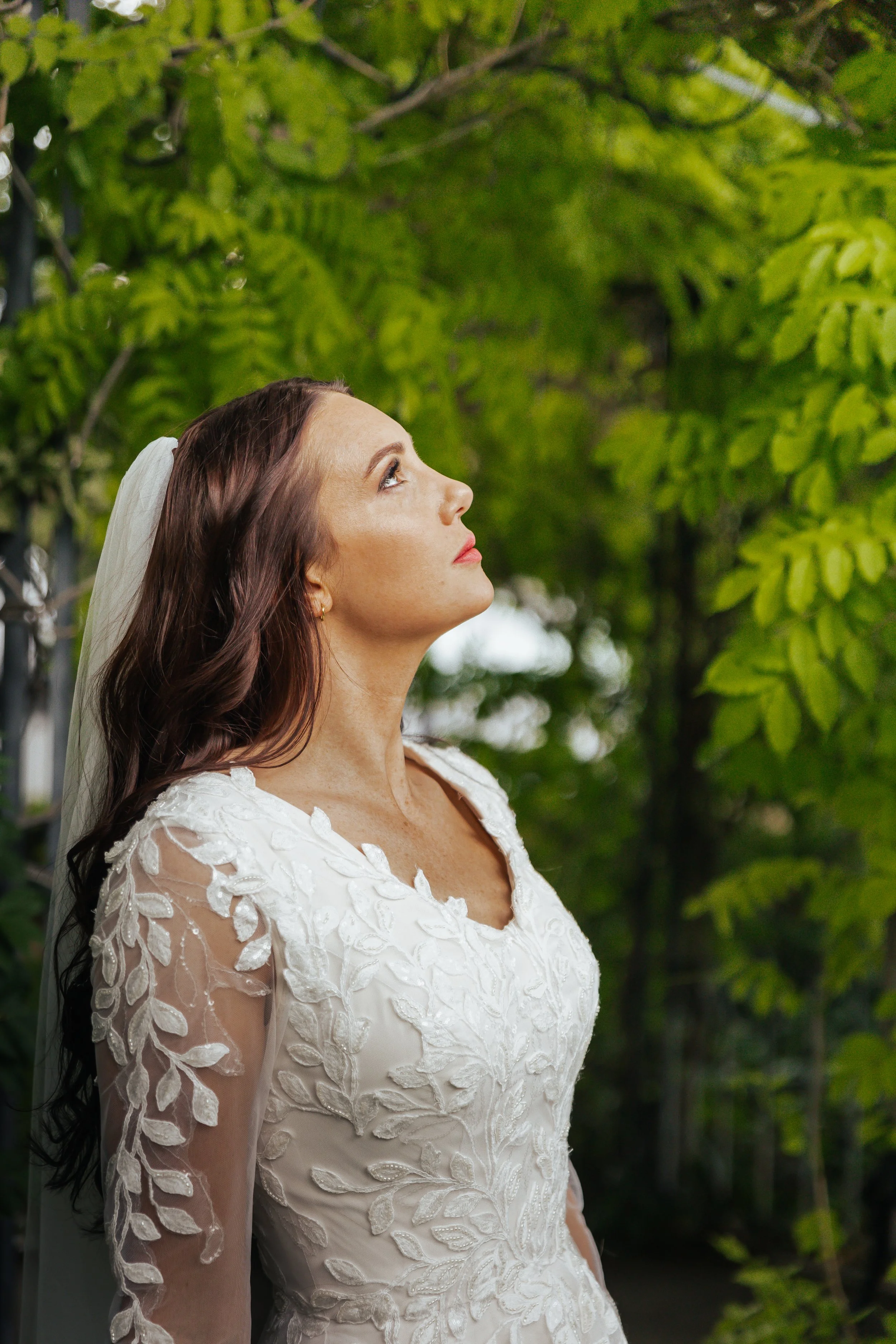 A bride with long dark hair in a white lace wedding dress with embroidered leaf patterns, standing outdoors against a background of green leaves, looking up.