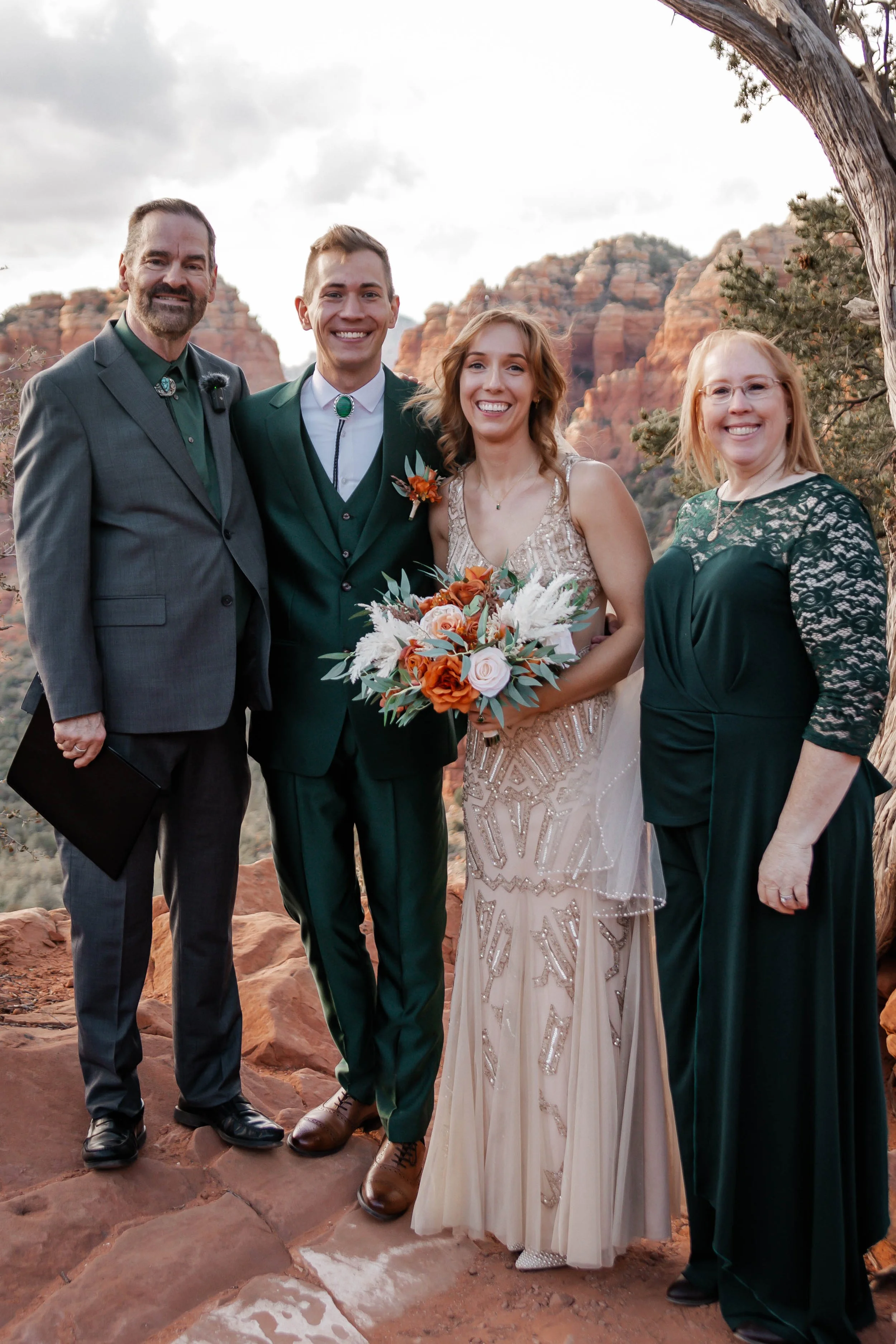 A wedding party standing outdoors, with a red rock canyon background, including a man in a gray suit, a man in a green suit, a woman in a cream dress holding a bouquet, and a woman in a dark green dress