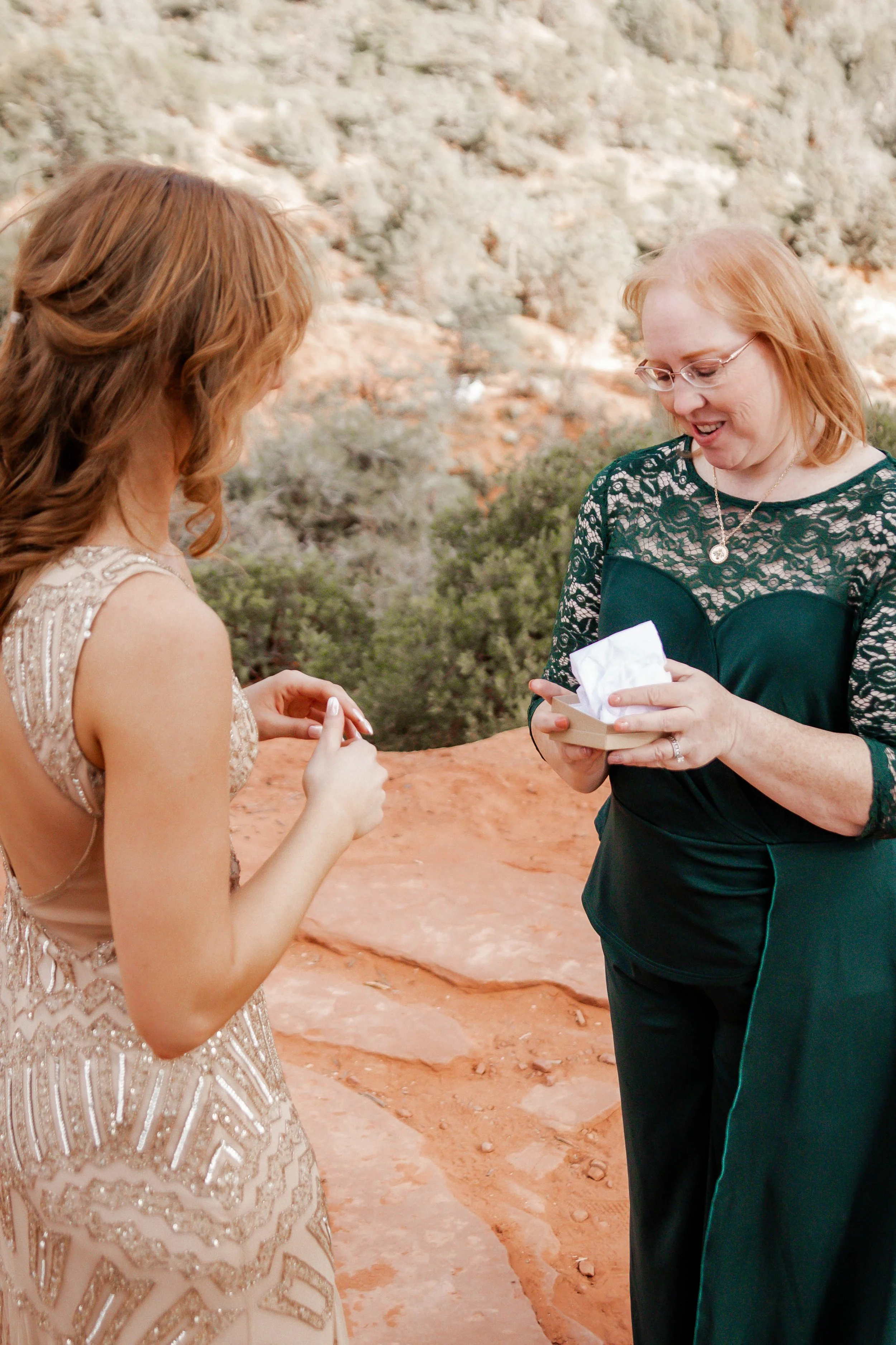 A woman in a black lace top and glasses is surprised as she receives a small gift box from a young woman in a beaded dress in a desert landscape.