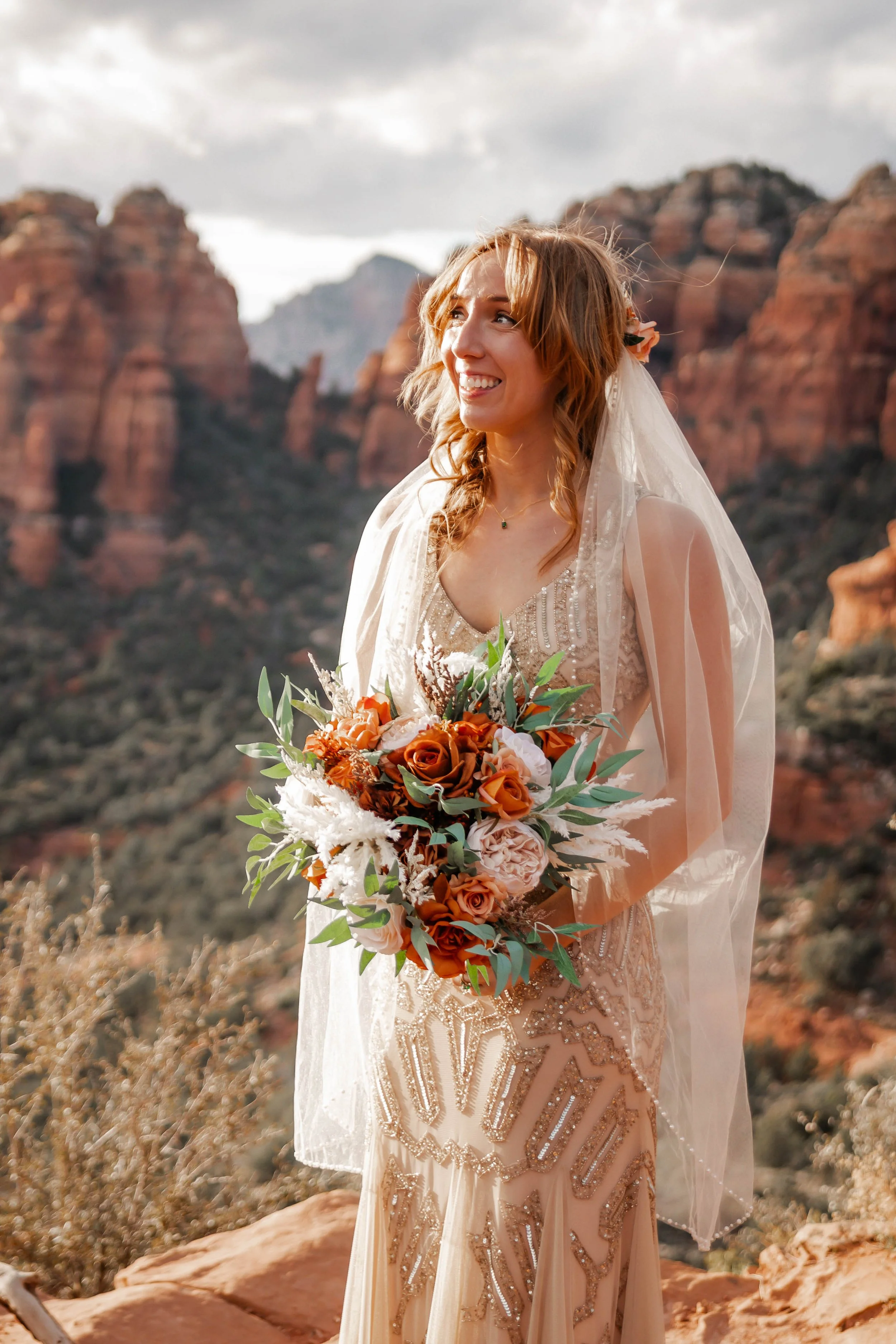 A woman in a detailed gown holding a bouquet of flowers, standing outdoors with red rock formations in the background.