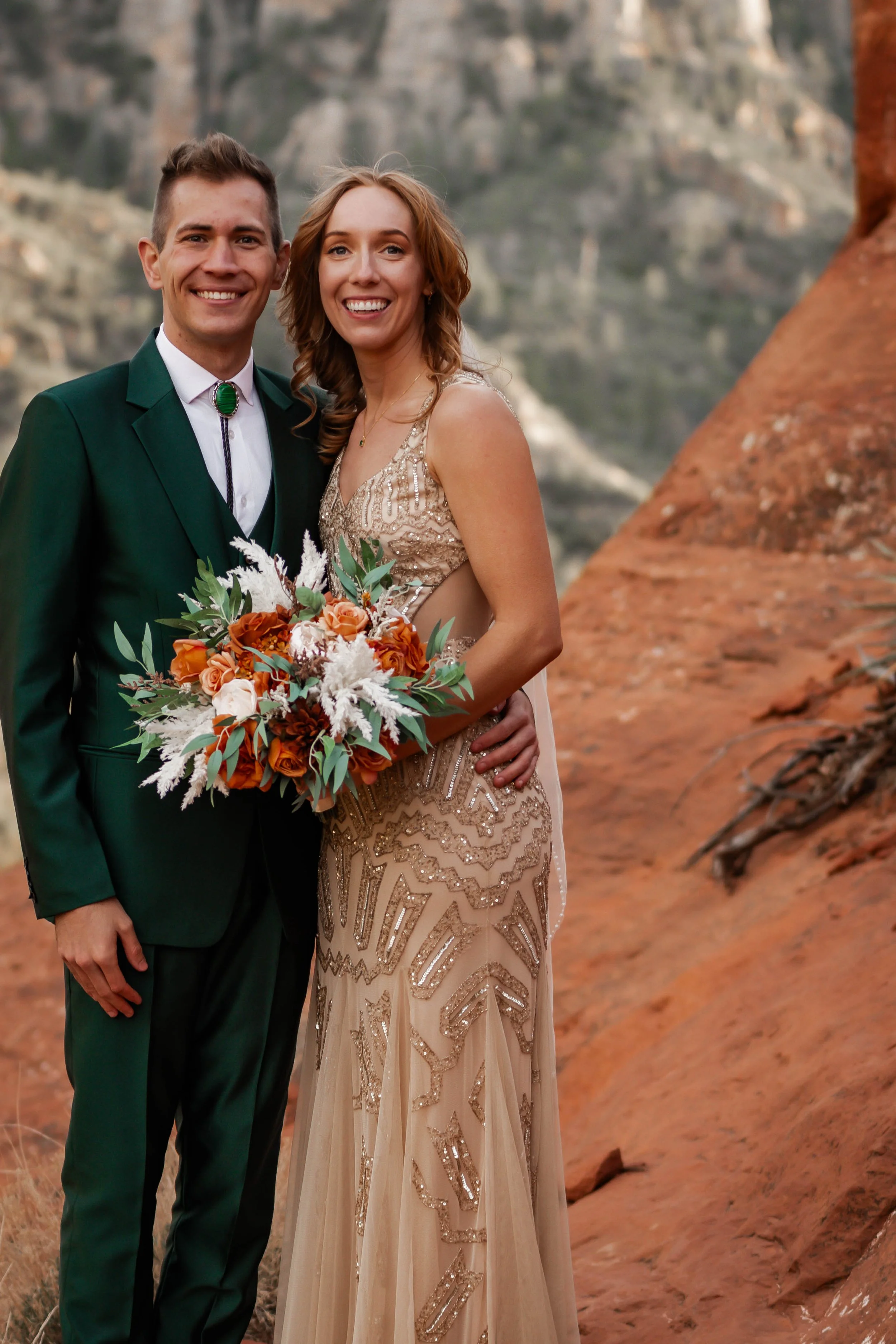 A happy bride and groom posing outdoors in front of red rocks, with the bride holding a bouquet of orange, white, and brown flowers, both smiling at the camera.