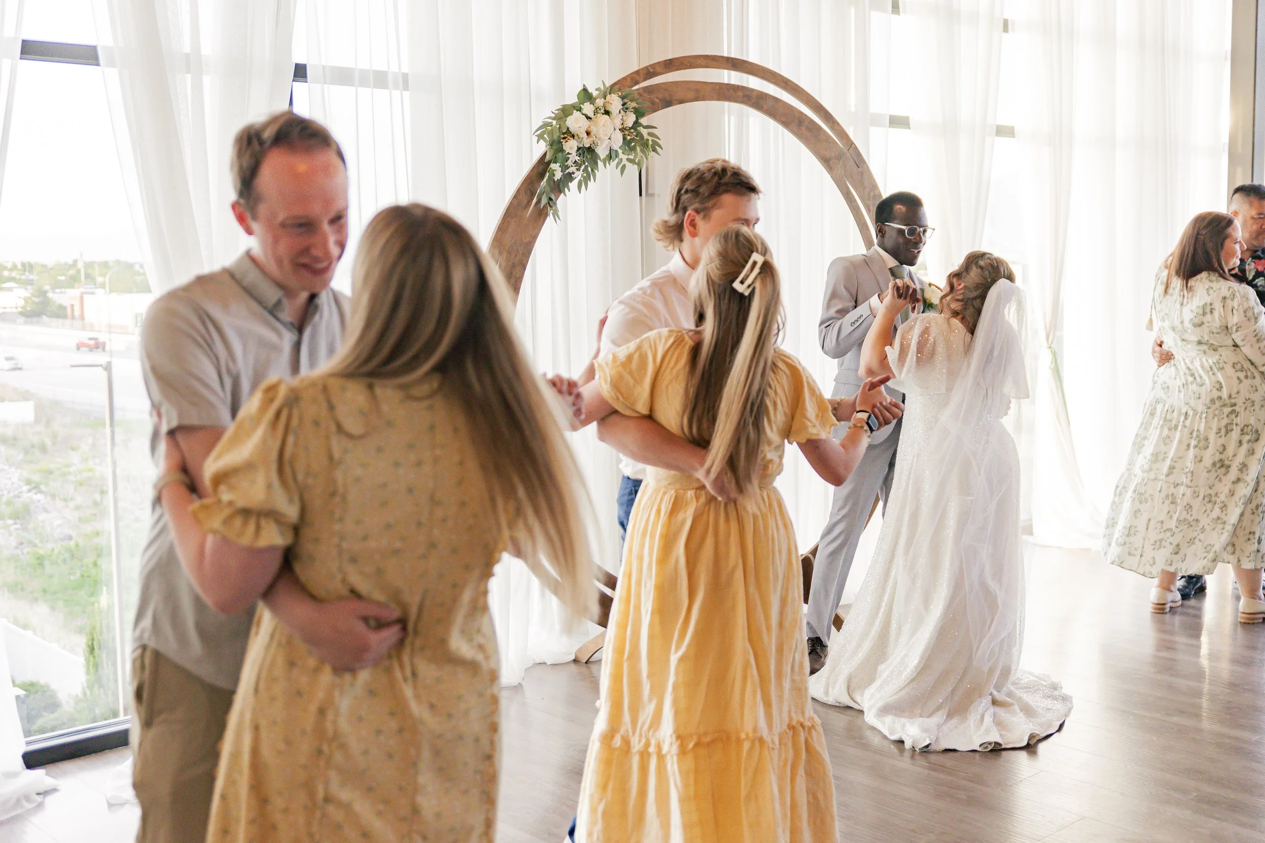 Couples dancing at a wedding reception, with a couple in the background dancing on a dance floor with a wooden arch decorated with flowers.