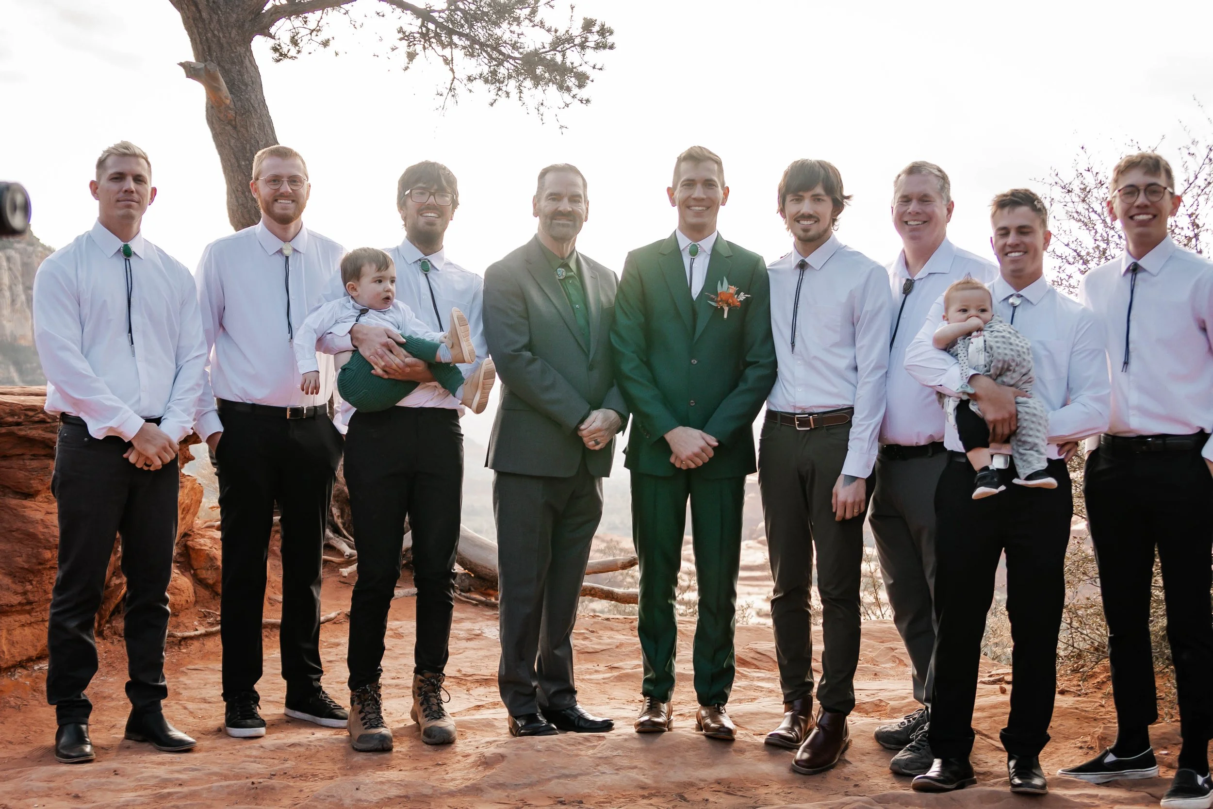 Group of men, including two in suits, standing outdoors on a sunny day, with a tree and rocky ground in the background.