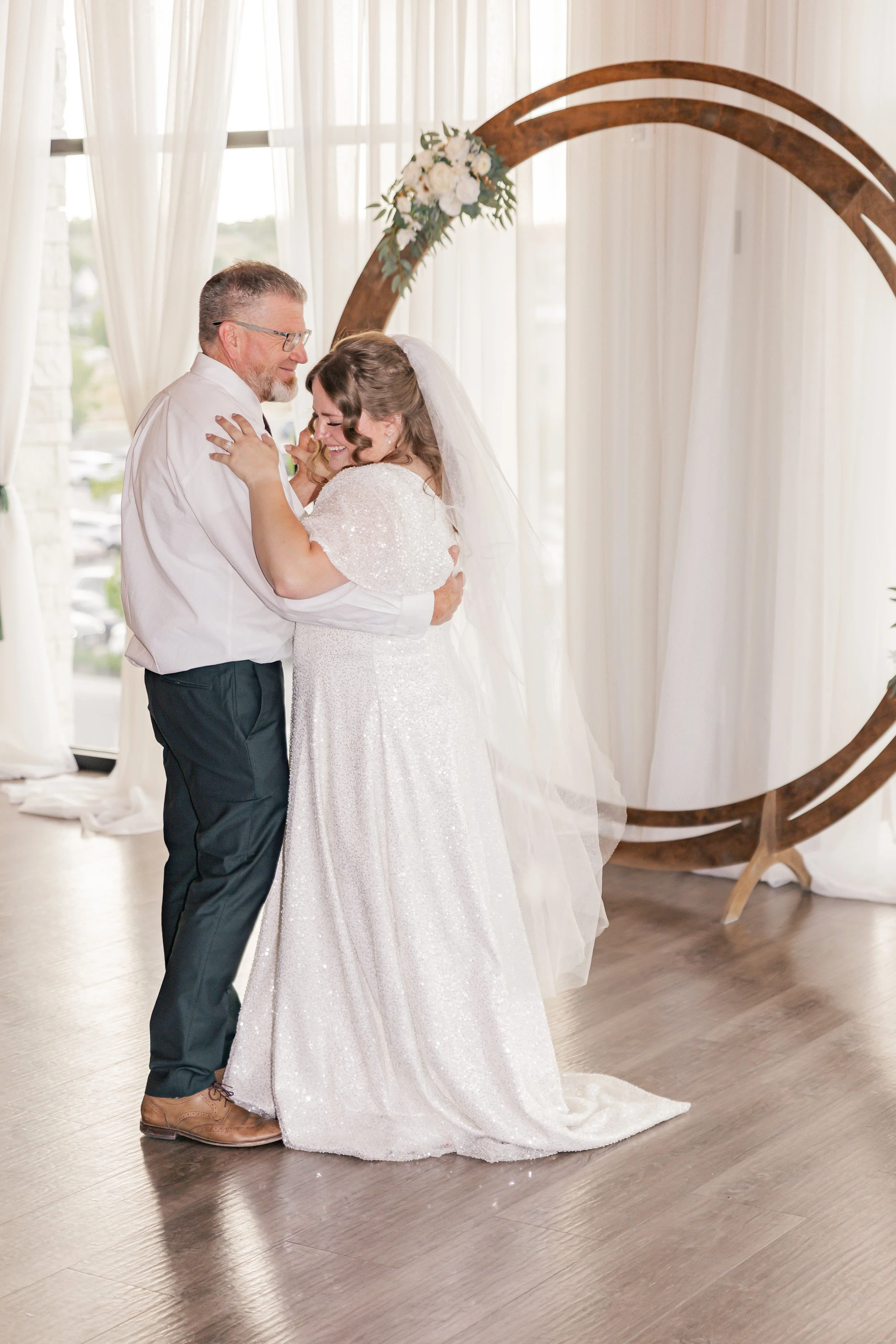 A bride and an older man, likely her father, sharing a dance at her wedding inside a venue with natural light and sheer curtains, with a decorative wooden circle with flowers in the background.