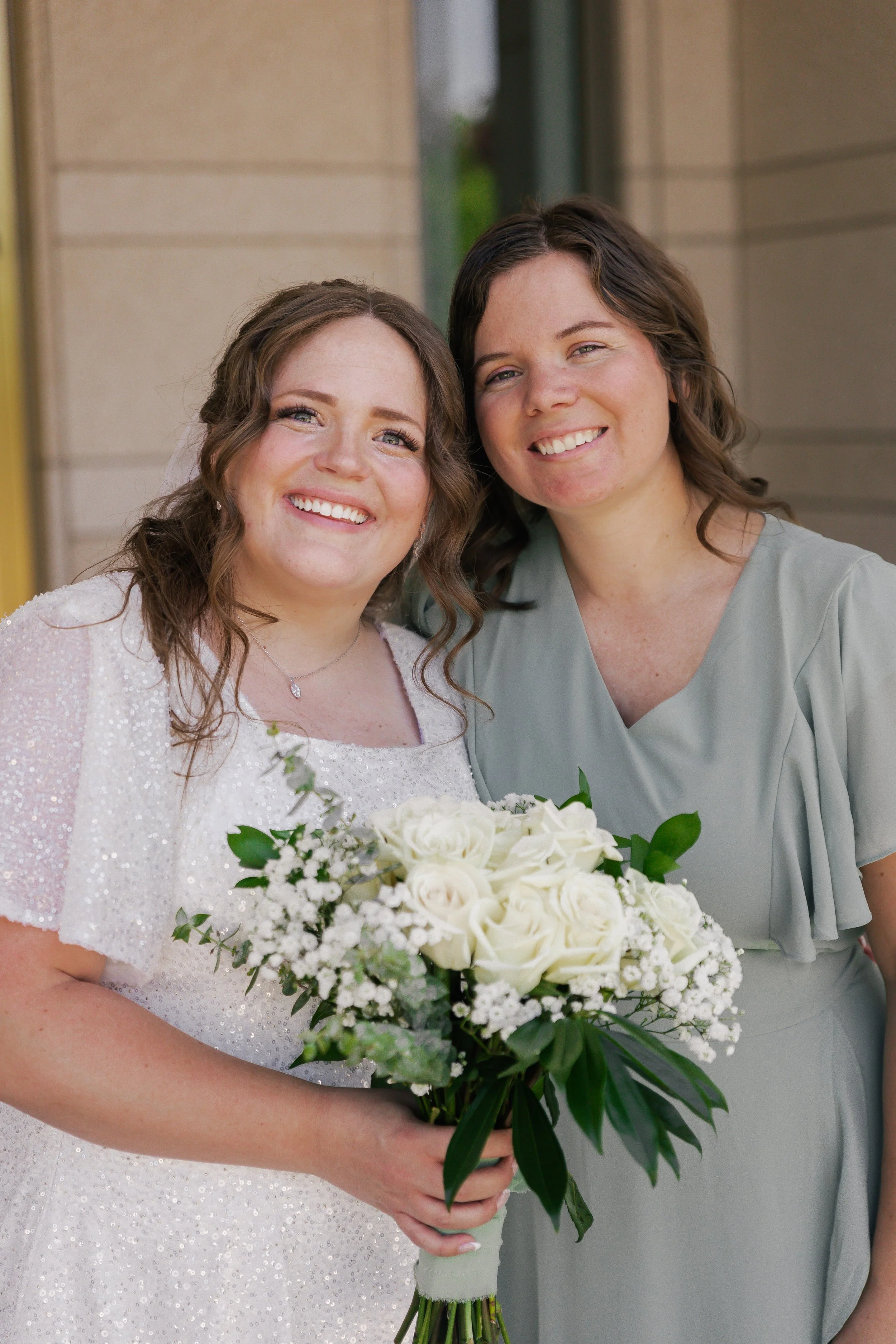 Two women, one in a wedding dress holding a bouquet of white roses, smiling outdoors.