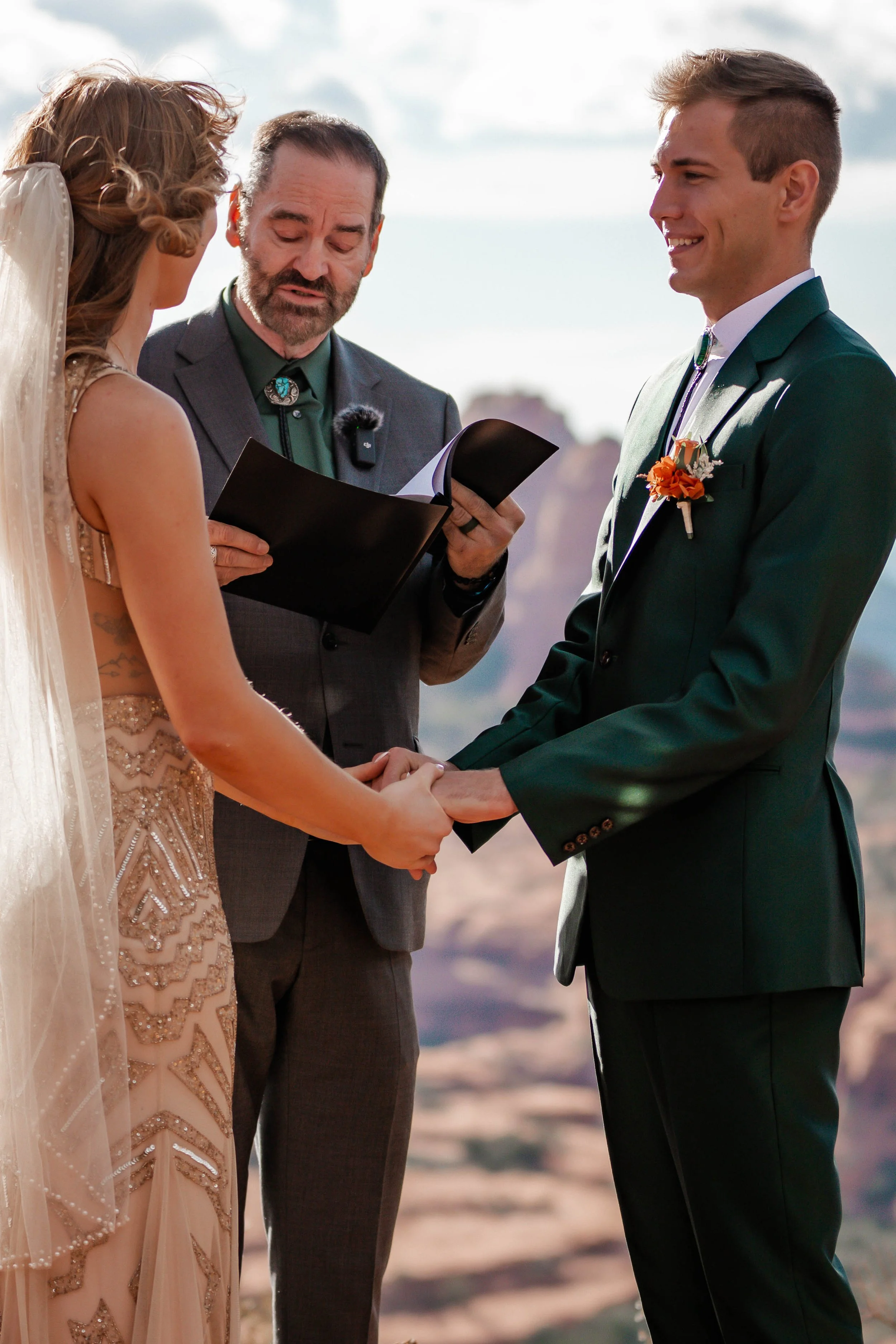 A couple getting married outdoors, holding hands, with a man officiating their ceremony, under a bright sky with a desert landscape in the background.