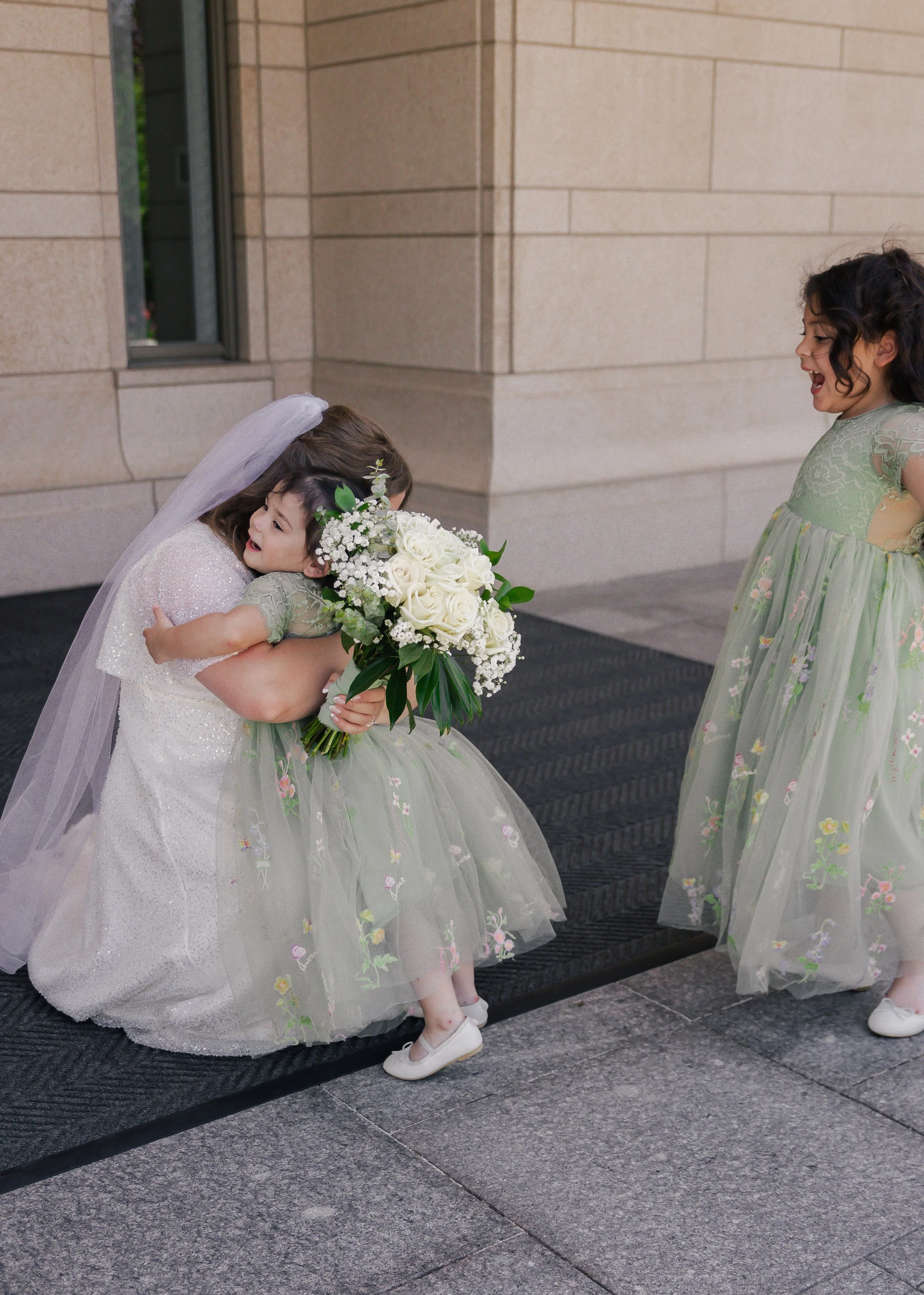 A bride hugging a young girl holding a bouquet of white roses and baby's breath outside a building, with another young girl standing nearby.