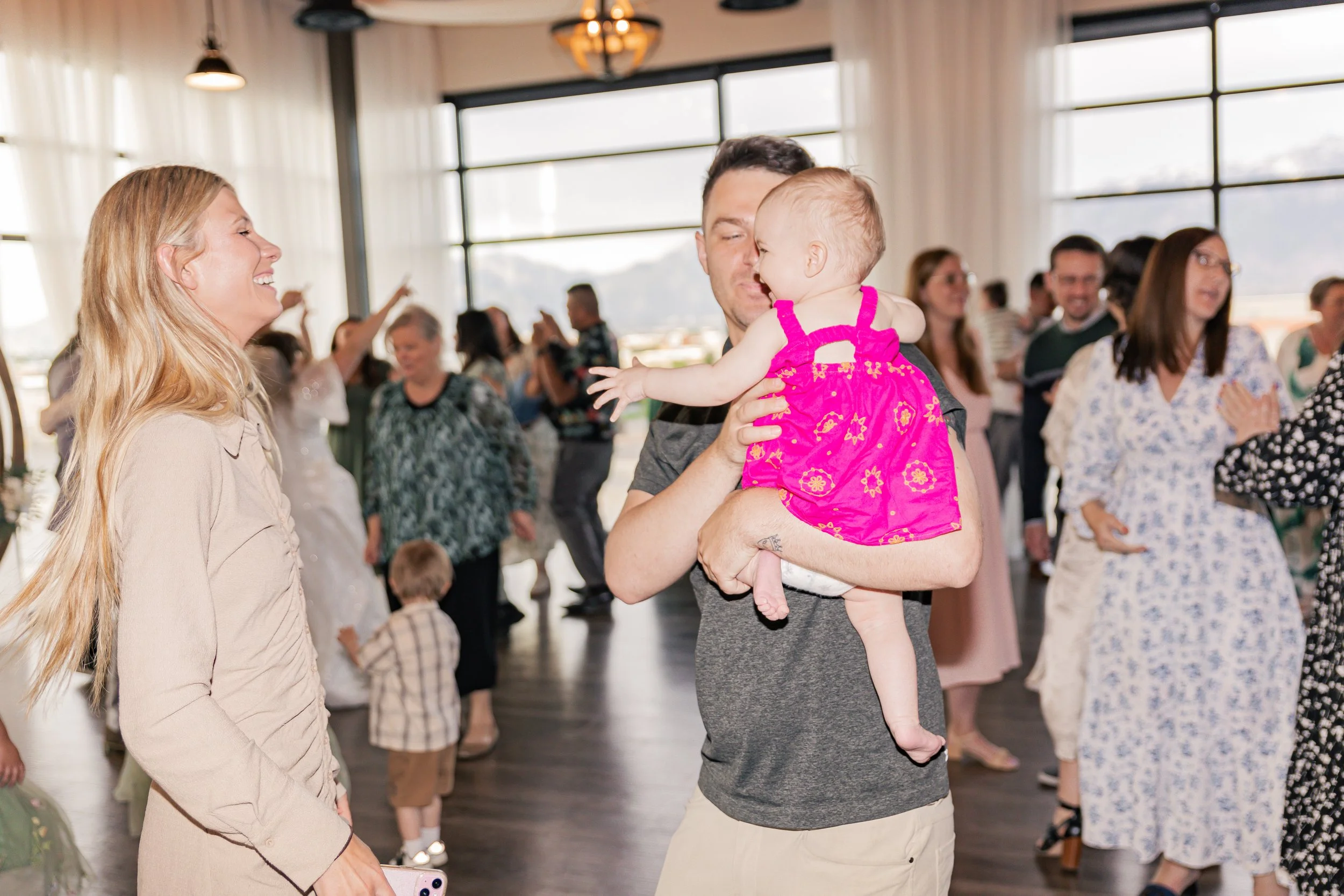 A man holding a smiling baby girl in a pink dress while dancing at a party or celebration with other people in the background.