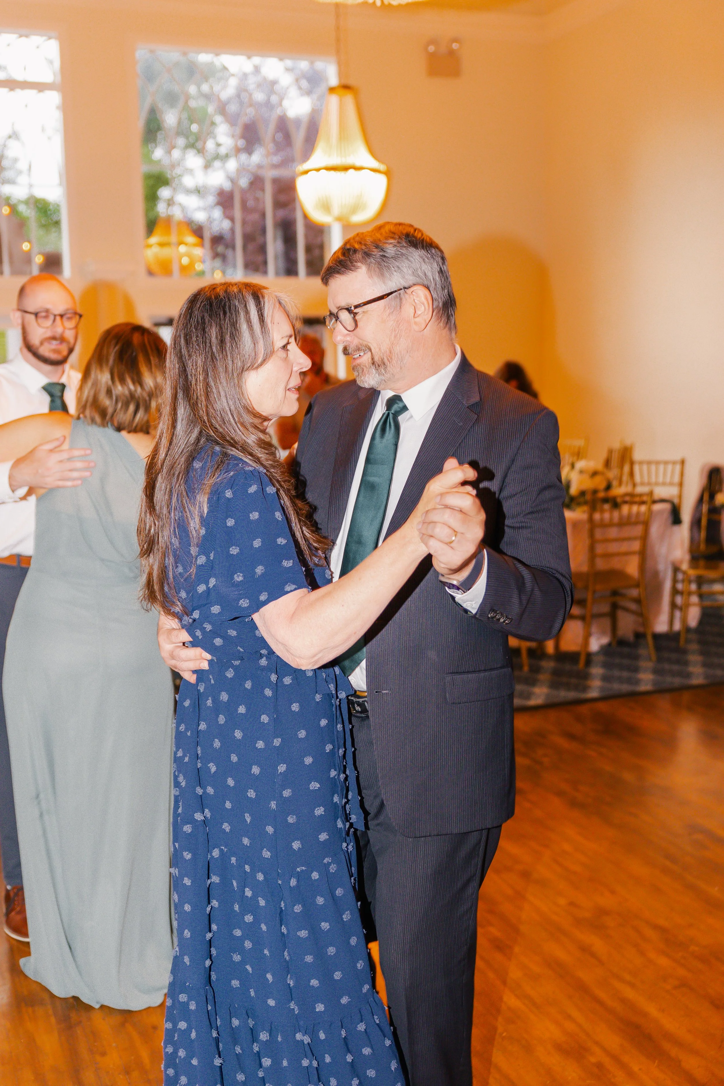 A couple dancing at a social gathering in a warmly lit room, with others dancing and socializing in the background.