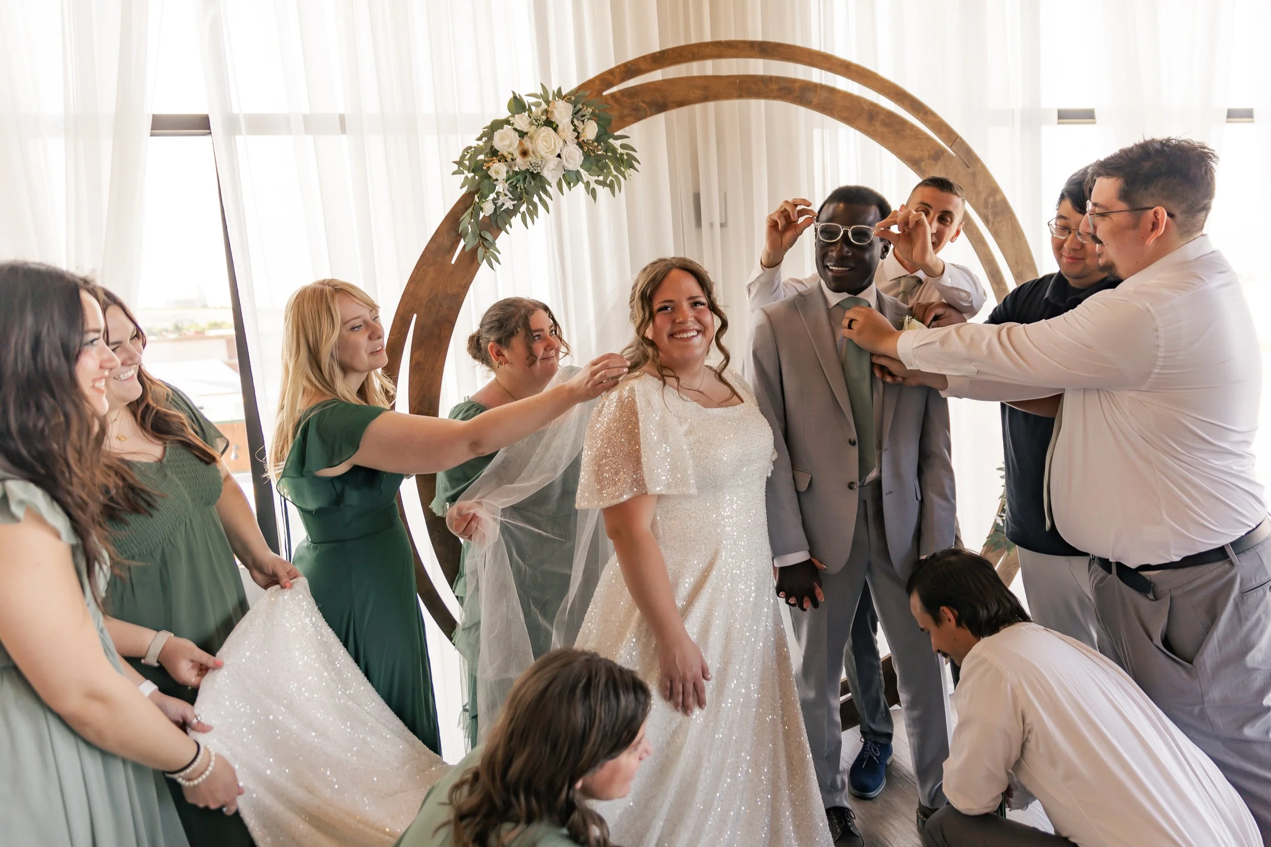 Bride in a white wedding dress surrounded by friends, with a decorative wooden arch and floral arrangement in the background.