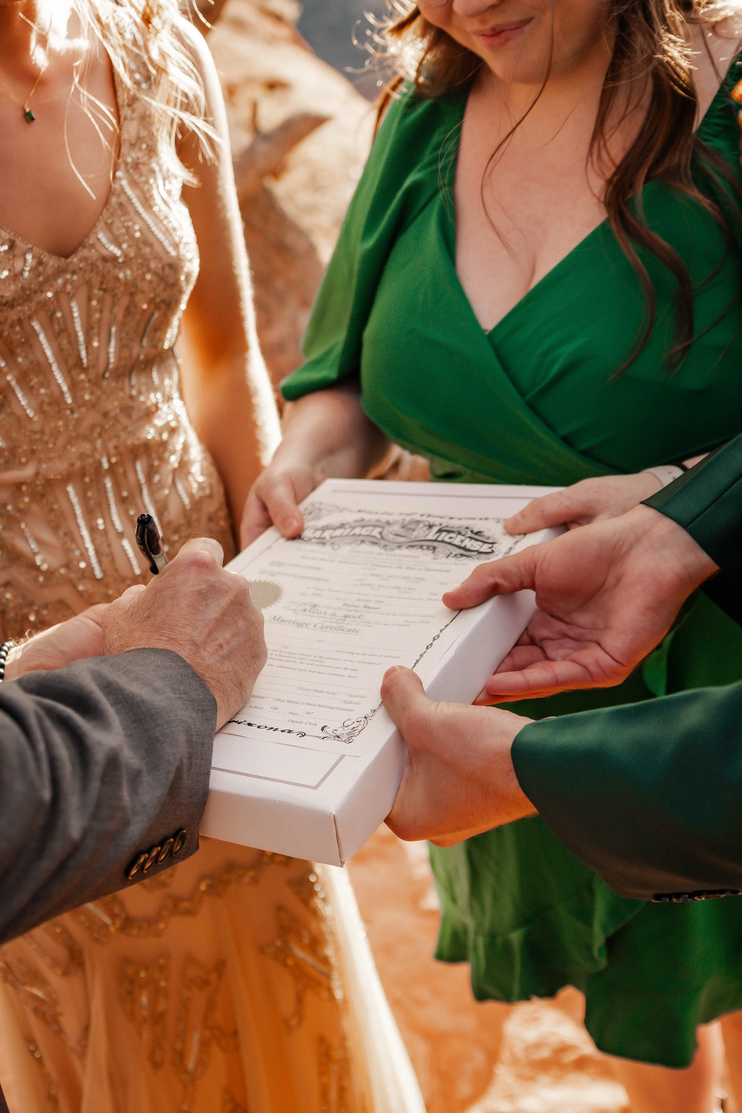 A wedding ceremony where a person signs a marriage license as the officiant holds a document and another person is holding a box containing the license. The bride is wearing a light pink dress with gold embellishments, and another woman beside her is