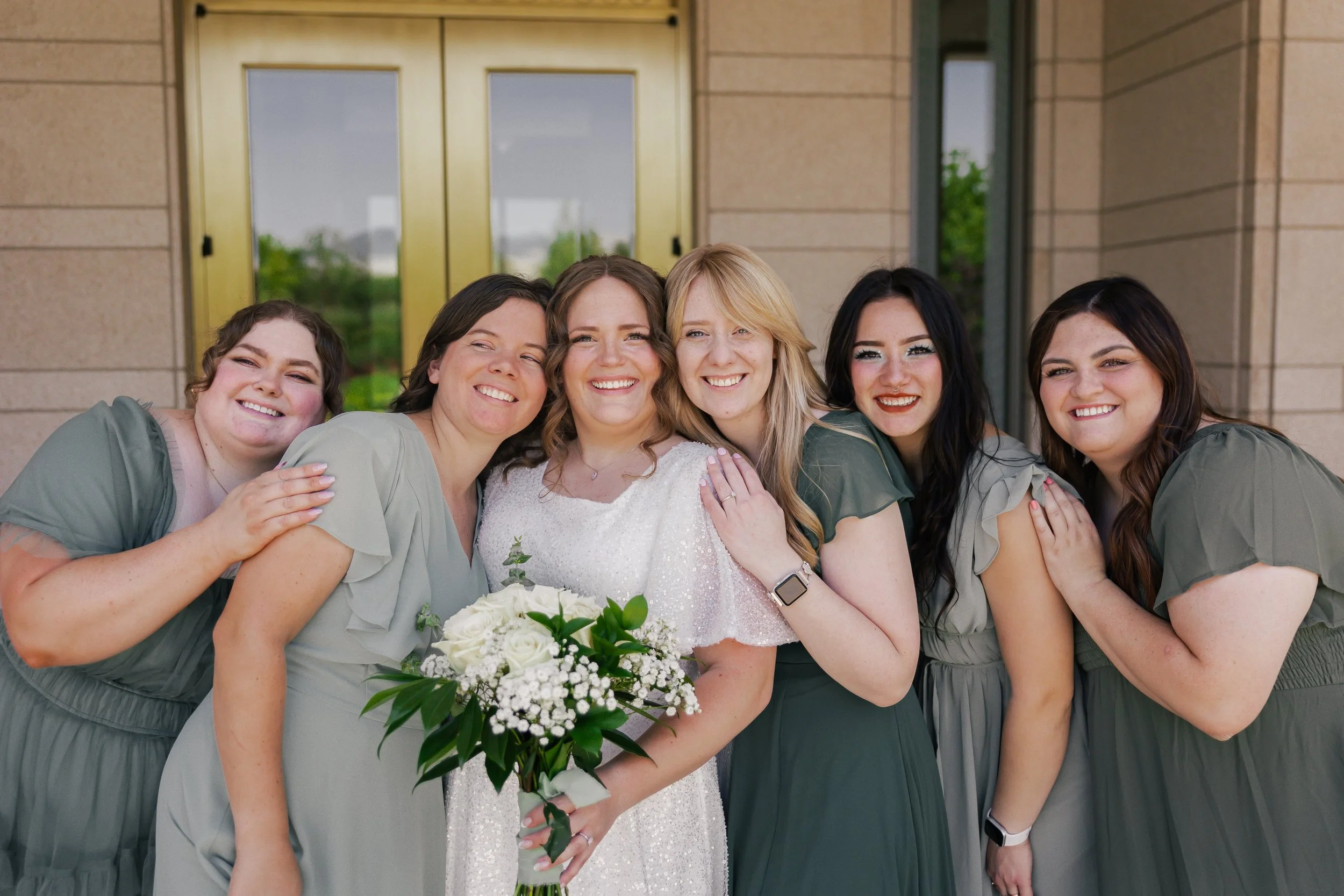 A group of women smiling, dressed in green and gray dresses, standing outside a building with a woman in a white dress holding a bouquet of flowers, likely at a wedding or special event.