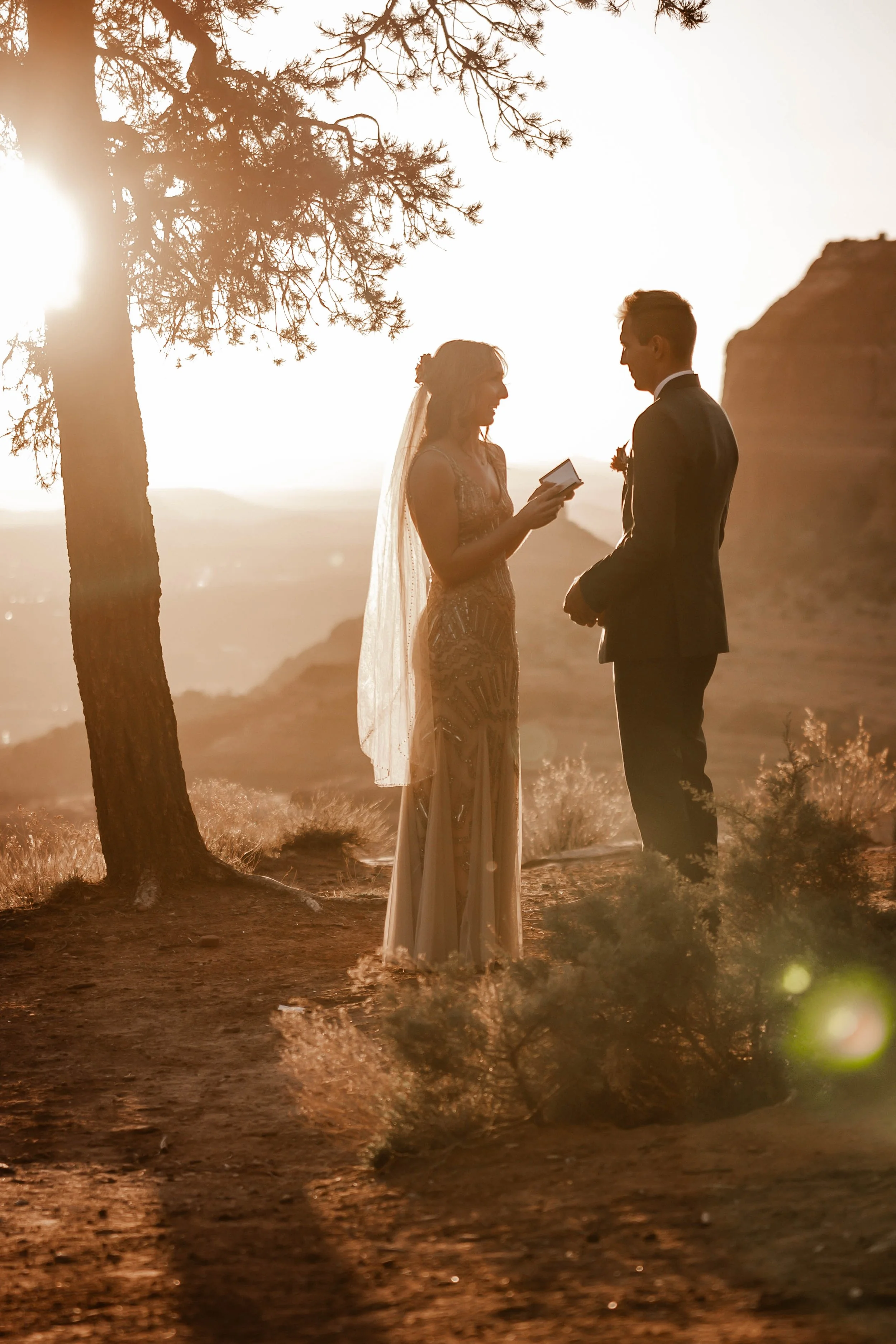 A bride and groom exchanging vows outdoors during sunset, with the bride reading from a book and the couple standing near a tree, with mountains in the background.