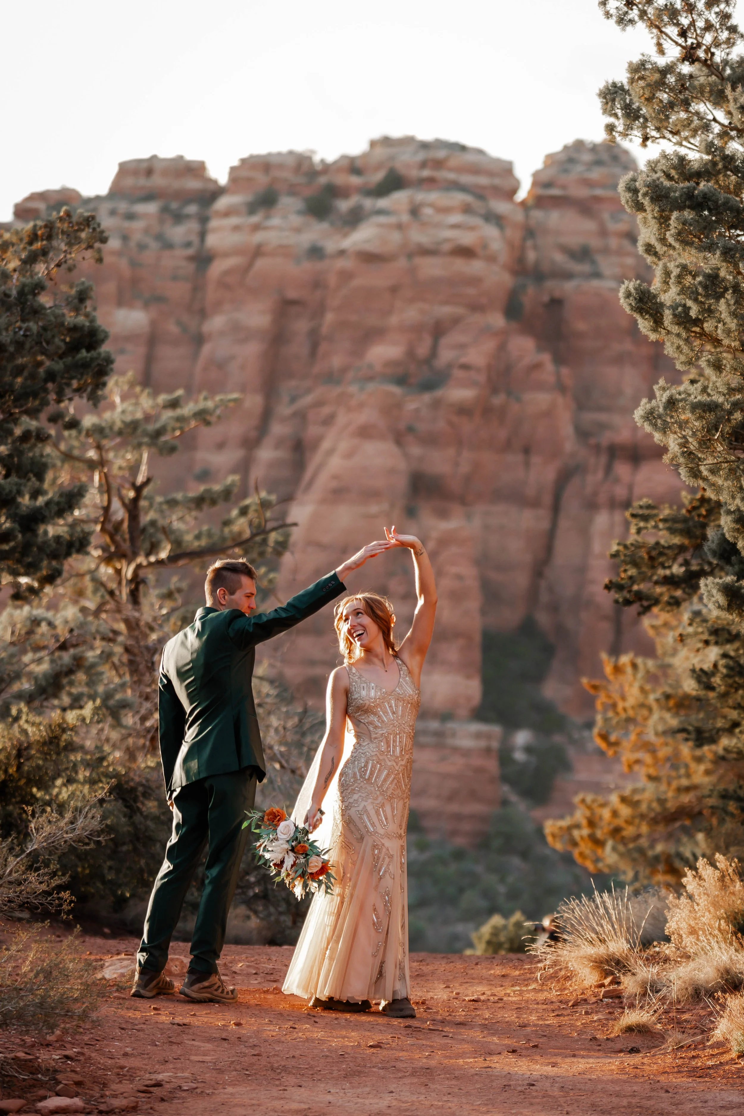 A couple dancing outdoors in front of a large rock formation, with the woman holding a bouquet of flowers and wearing a wedding dress, and the man wearing a dark suit, during sunset.