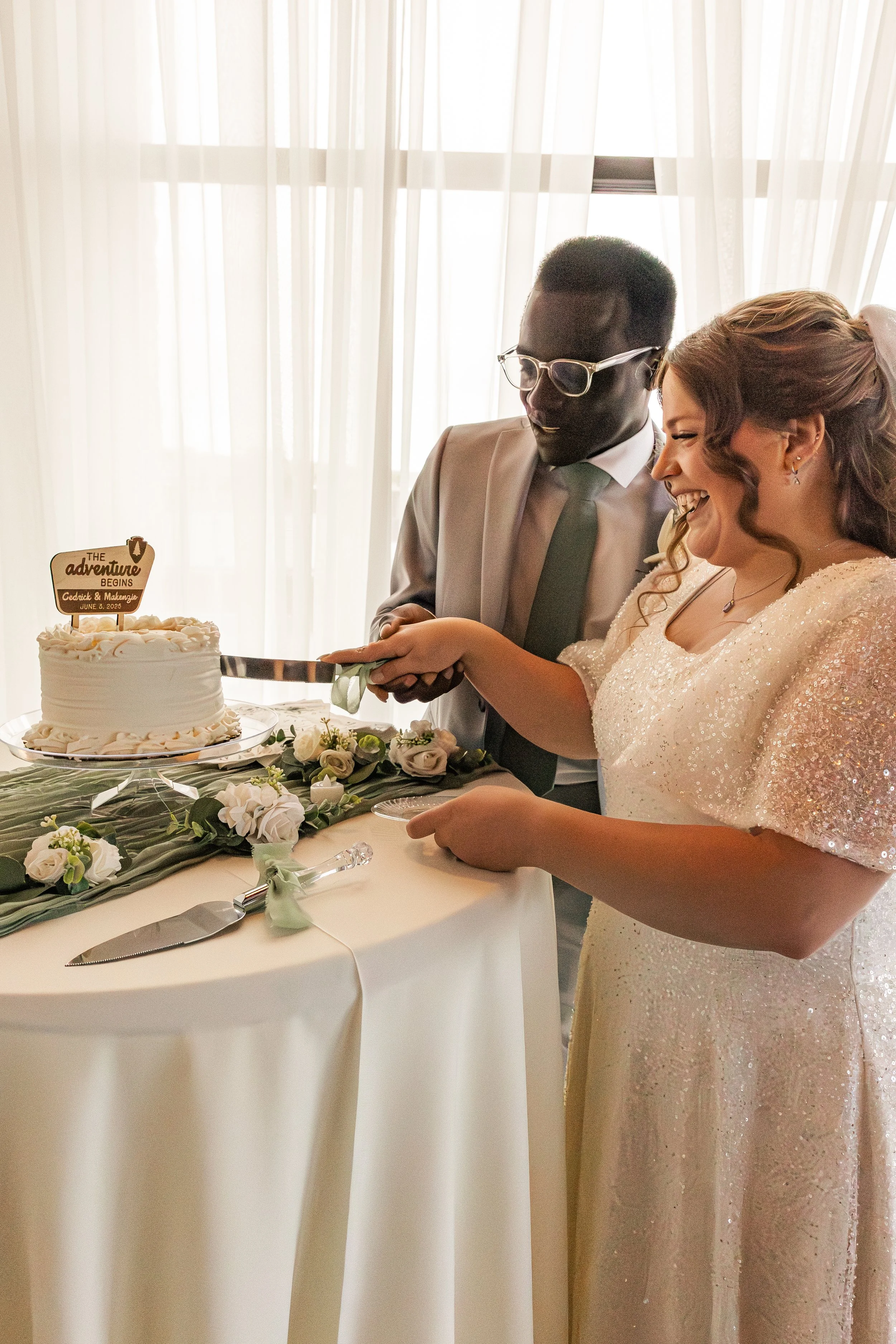 A bride and groom cutting wedding cake together, with the bride smiling and the groom holding a cake knife, standing in front of a white curtain.
