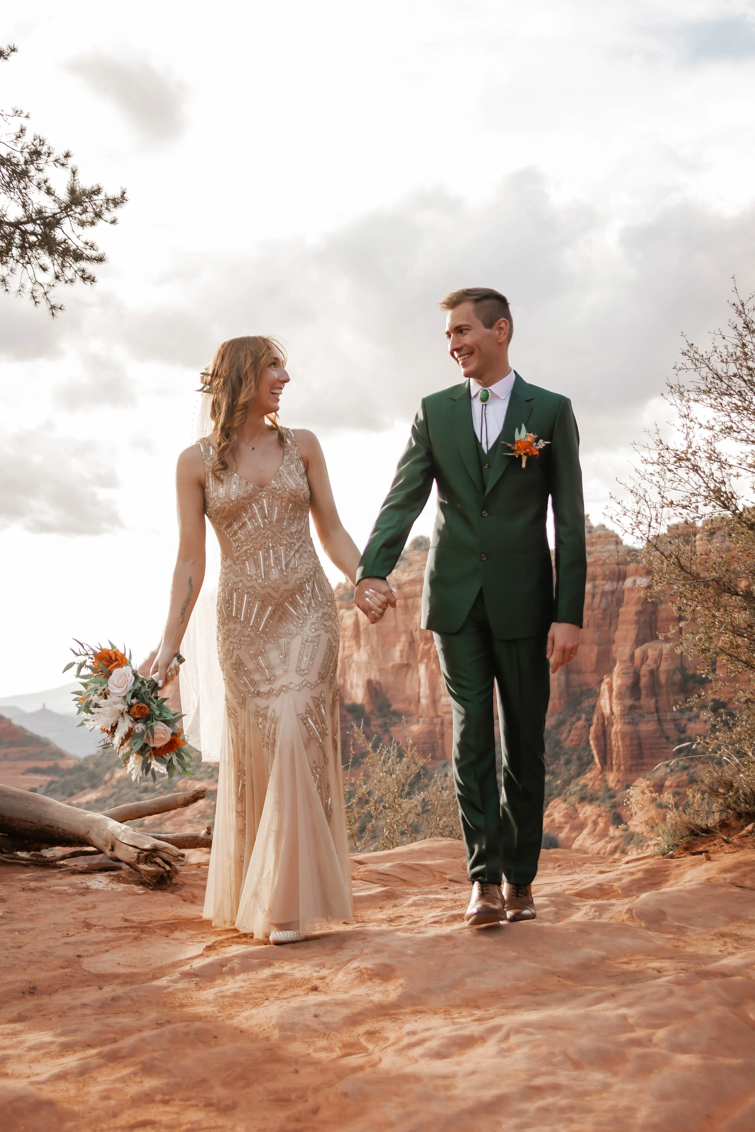 A bride and groom walking hand in hand on a rocky desert landscape with red rock formations in the background, smiling at each other.