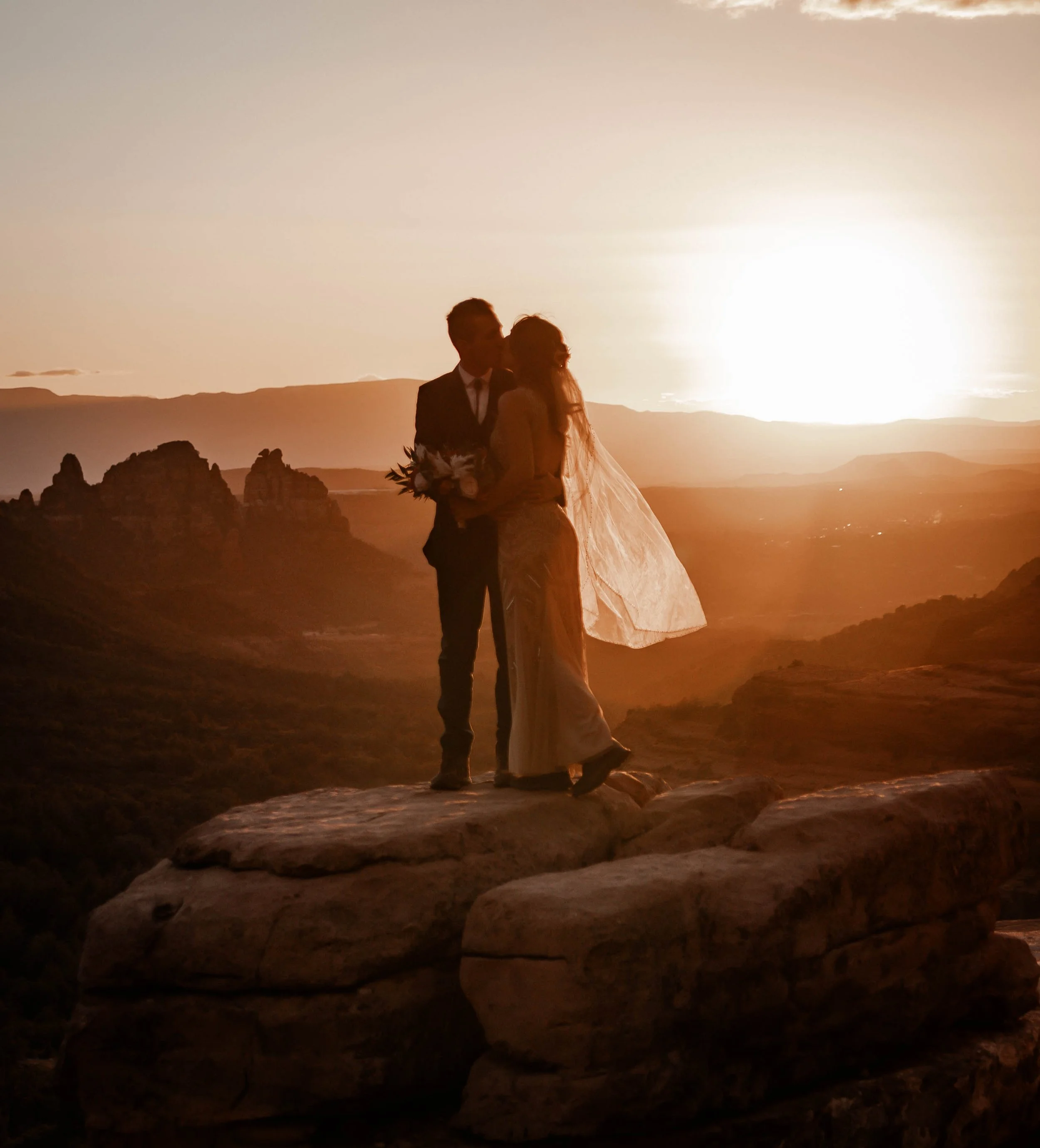 Silhouette of a bride and groom kissing on a rock during sunset with a scenic valley and mountains in the background.