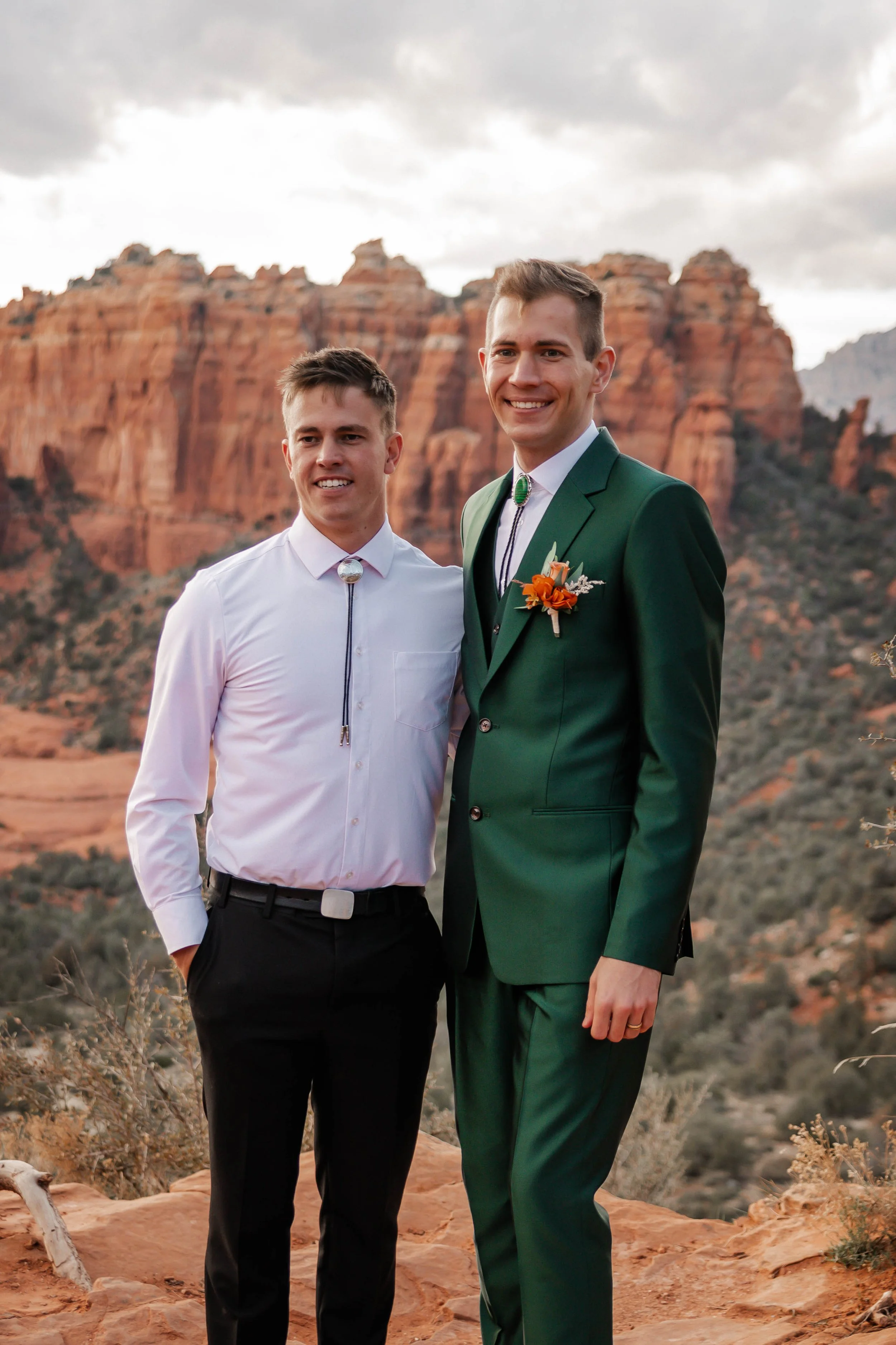 Two men, one in a dark green suit and the other in a white shirt and black pants, stand outdoors with red rock formations in the background.