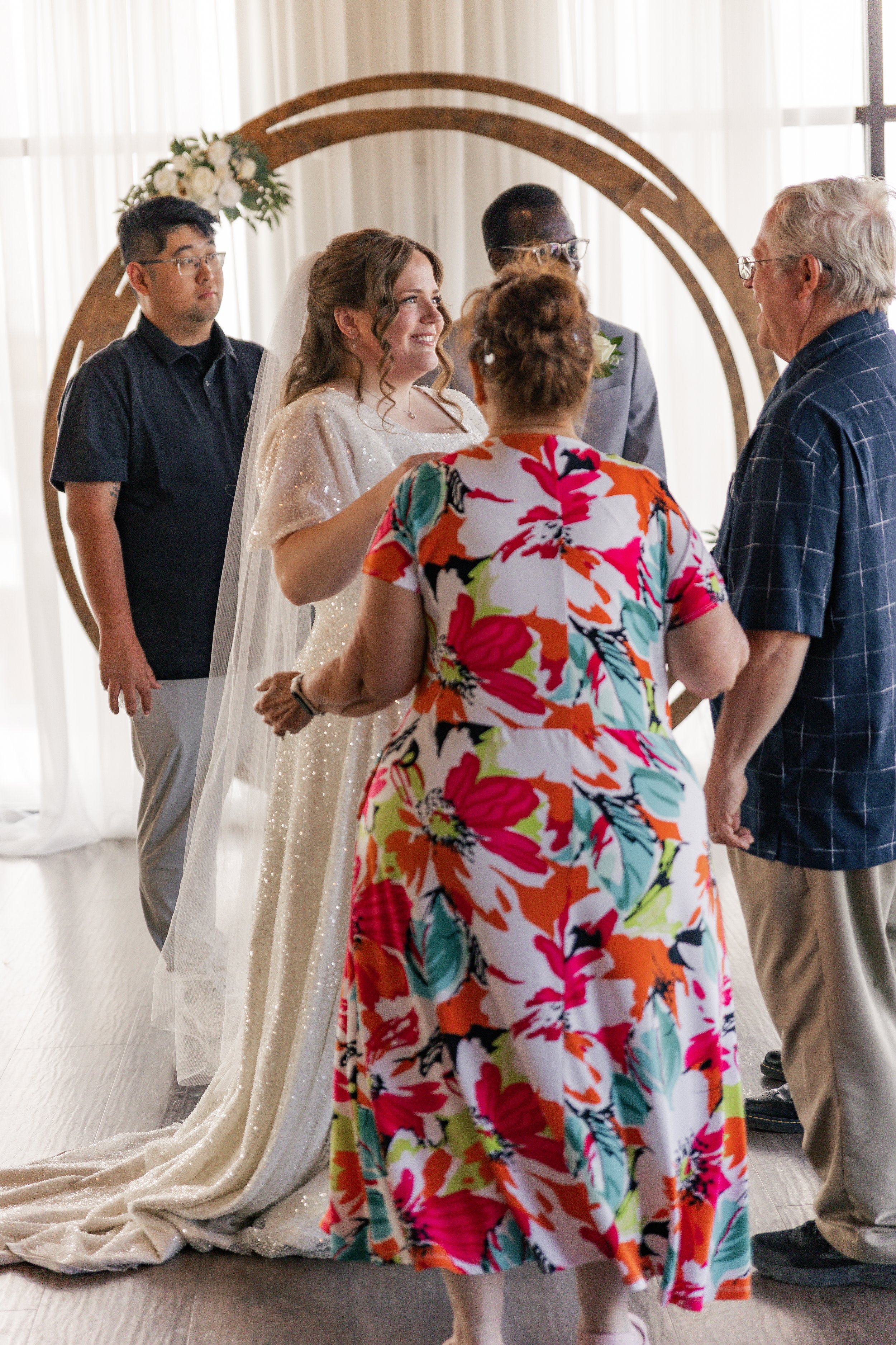 A couple getting married, standing in front of a wooden arch decorated with white flowers, with three people around them in a bright room with sheer curtains.