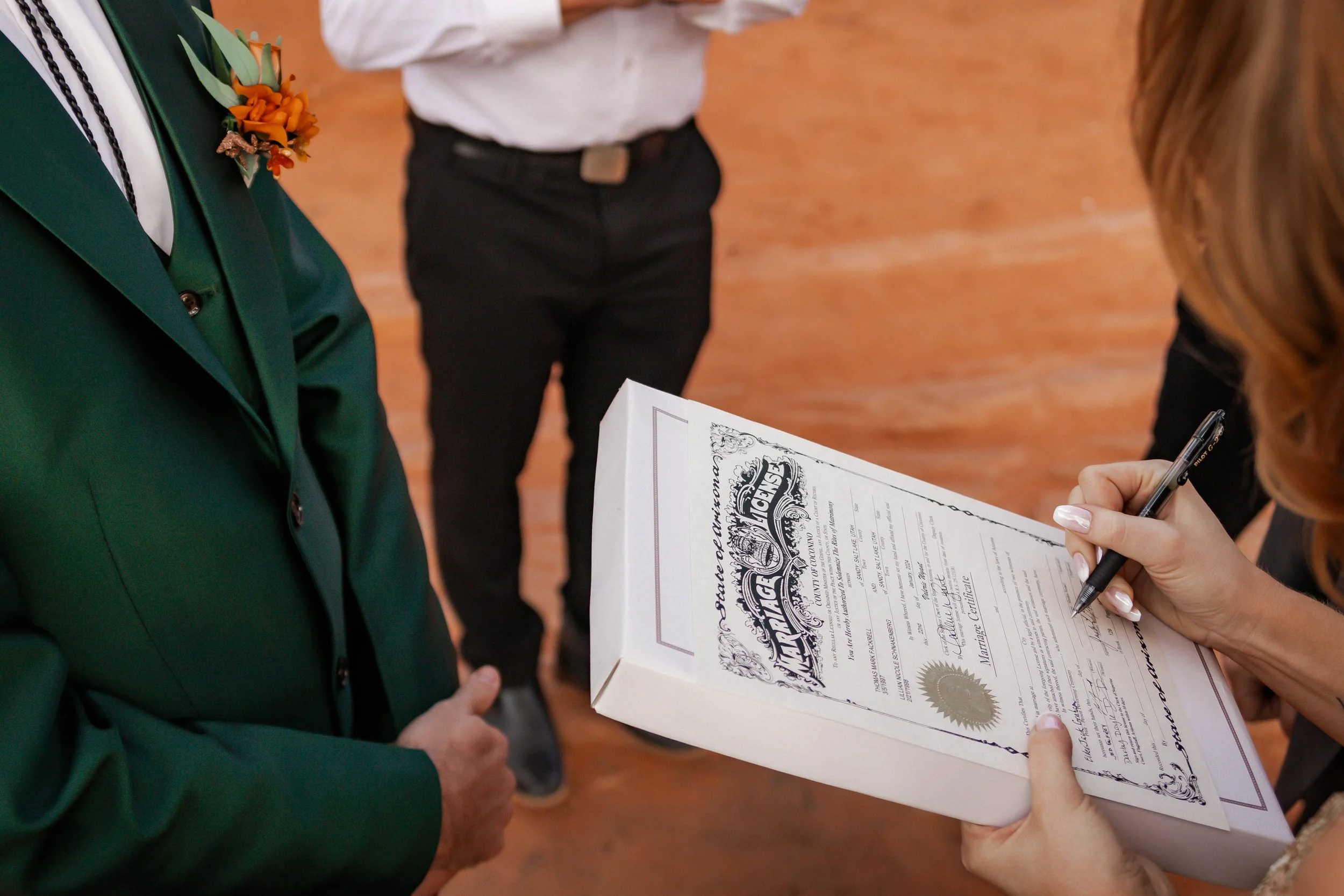 A person signing a marriage certificate during a wedding ceremony, with a groom standing nearby and another person holding the certificate.