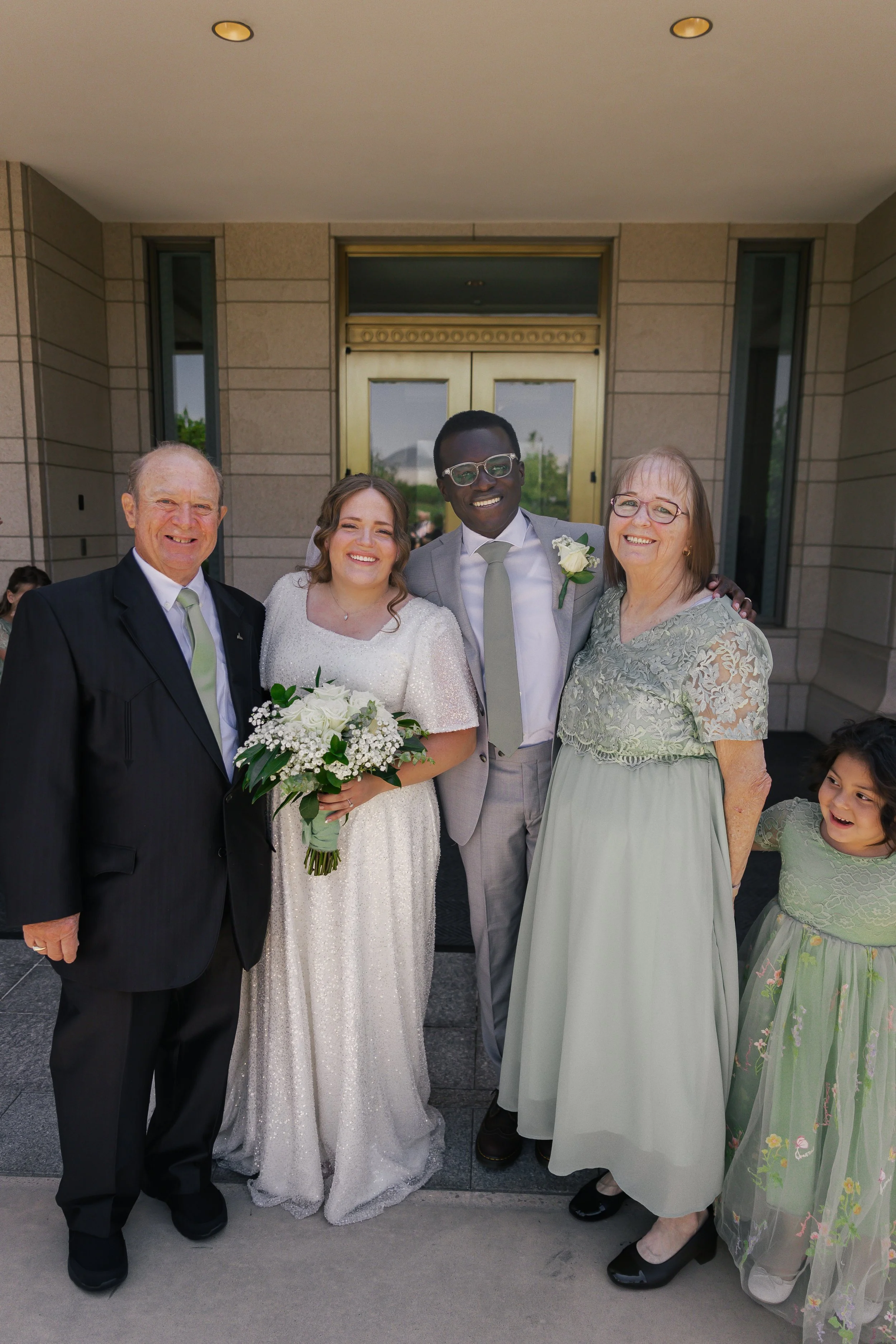 Group of five people, including a bride holding a bouquet, standing in front of a building with a gold door during a wedding celebration.