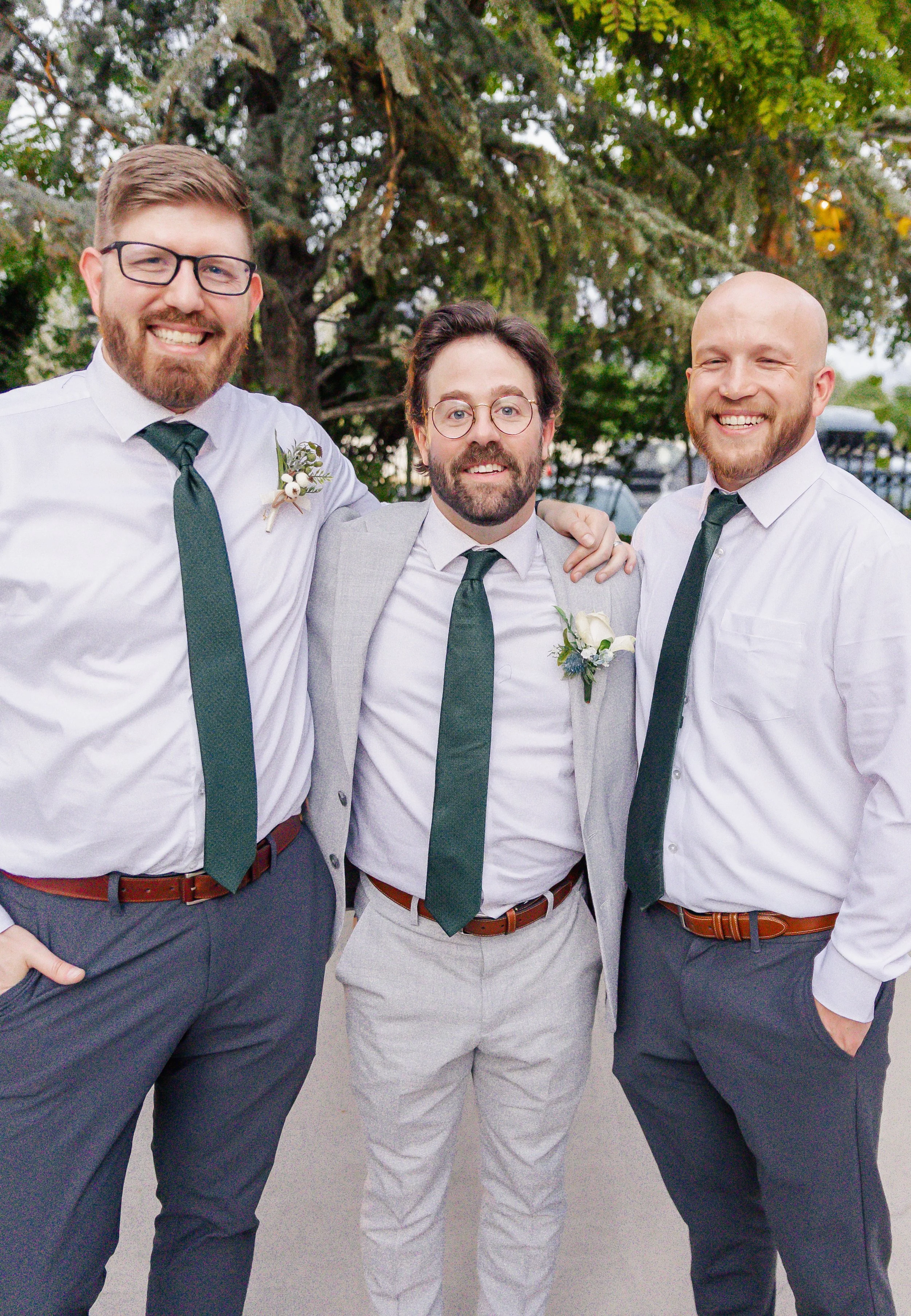Three men dressed in white shirts, dark green ties, and dress pants, standing outdoors in front of a large tree. They are smiling and have their arms around each other, celebrating a special occasion.