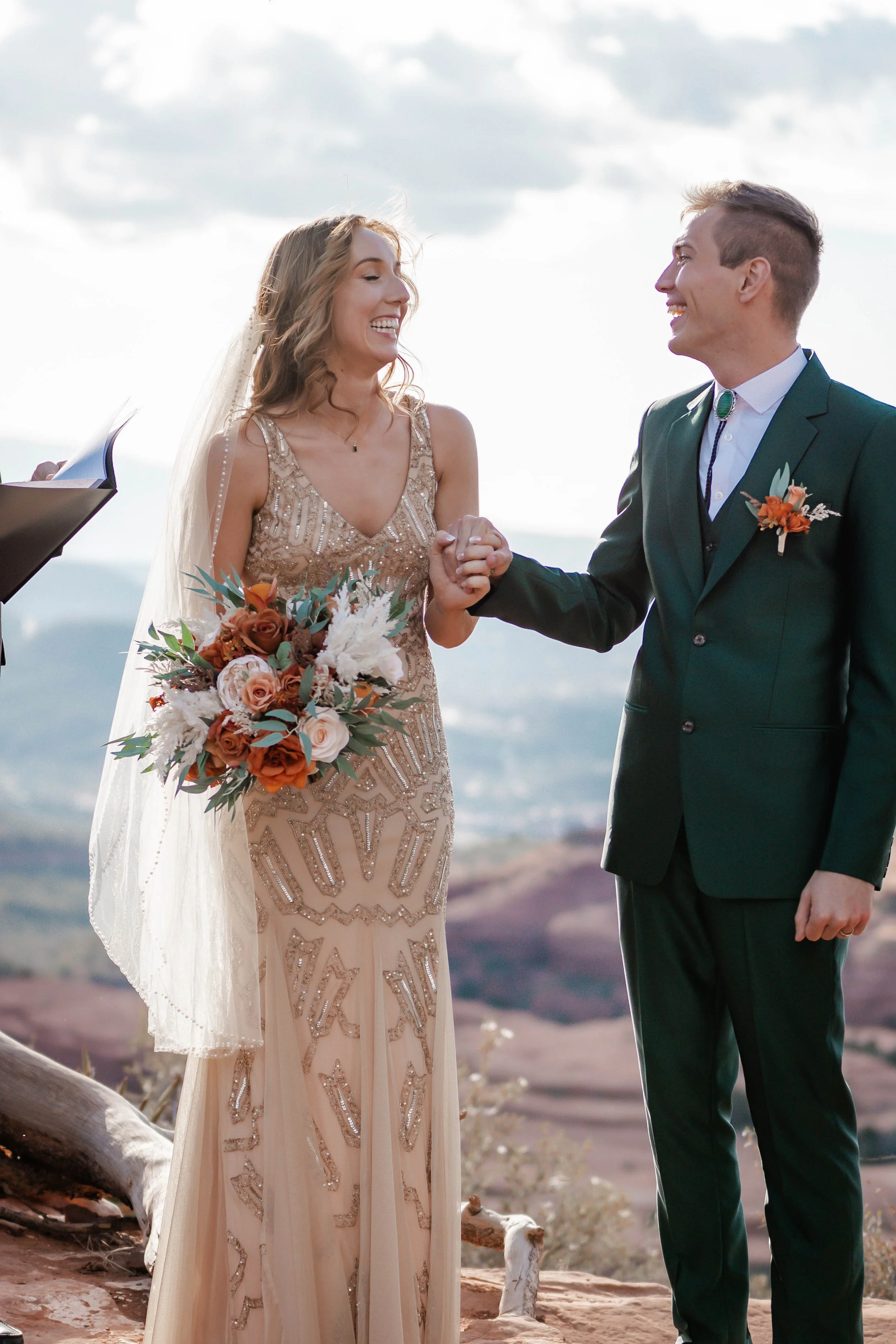 A bride in a beaded gown and a groom in a dark green suit holding hands during their wedding ceremony outdoors, with mountains and sky in the background.