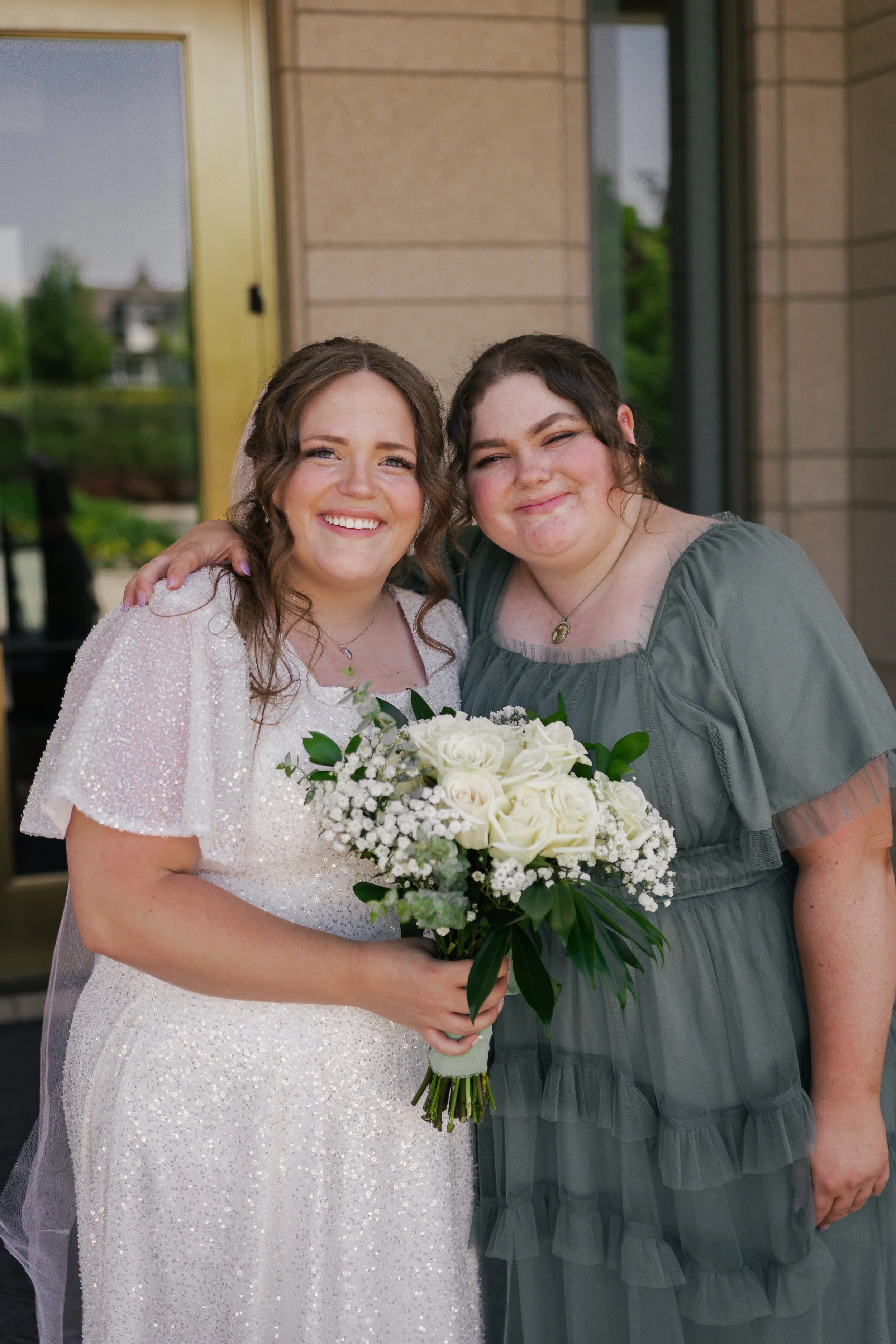 Two women smiling, one in a wedding dress holding a bouquet of white roses, the other in a green dress, standing outside a building.