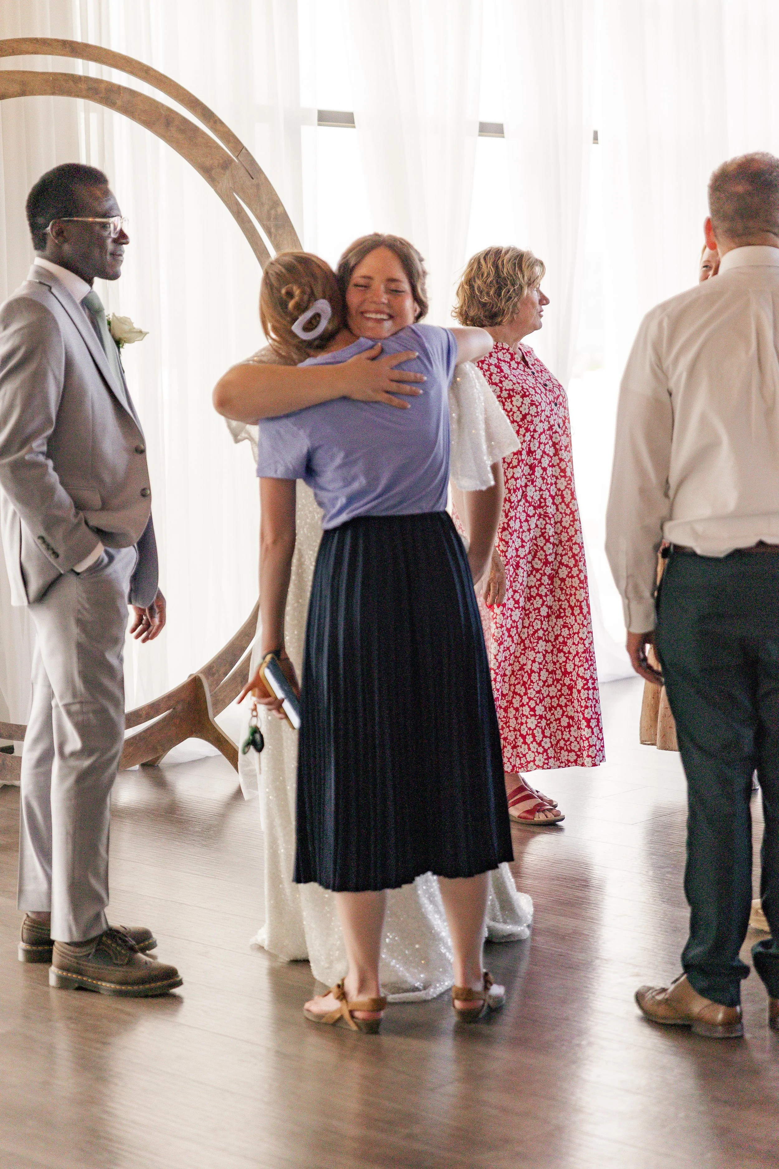 People hugging at a wedding or celebration indoors with bright curtains and wooden floor.