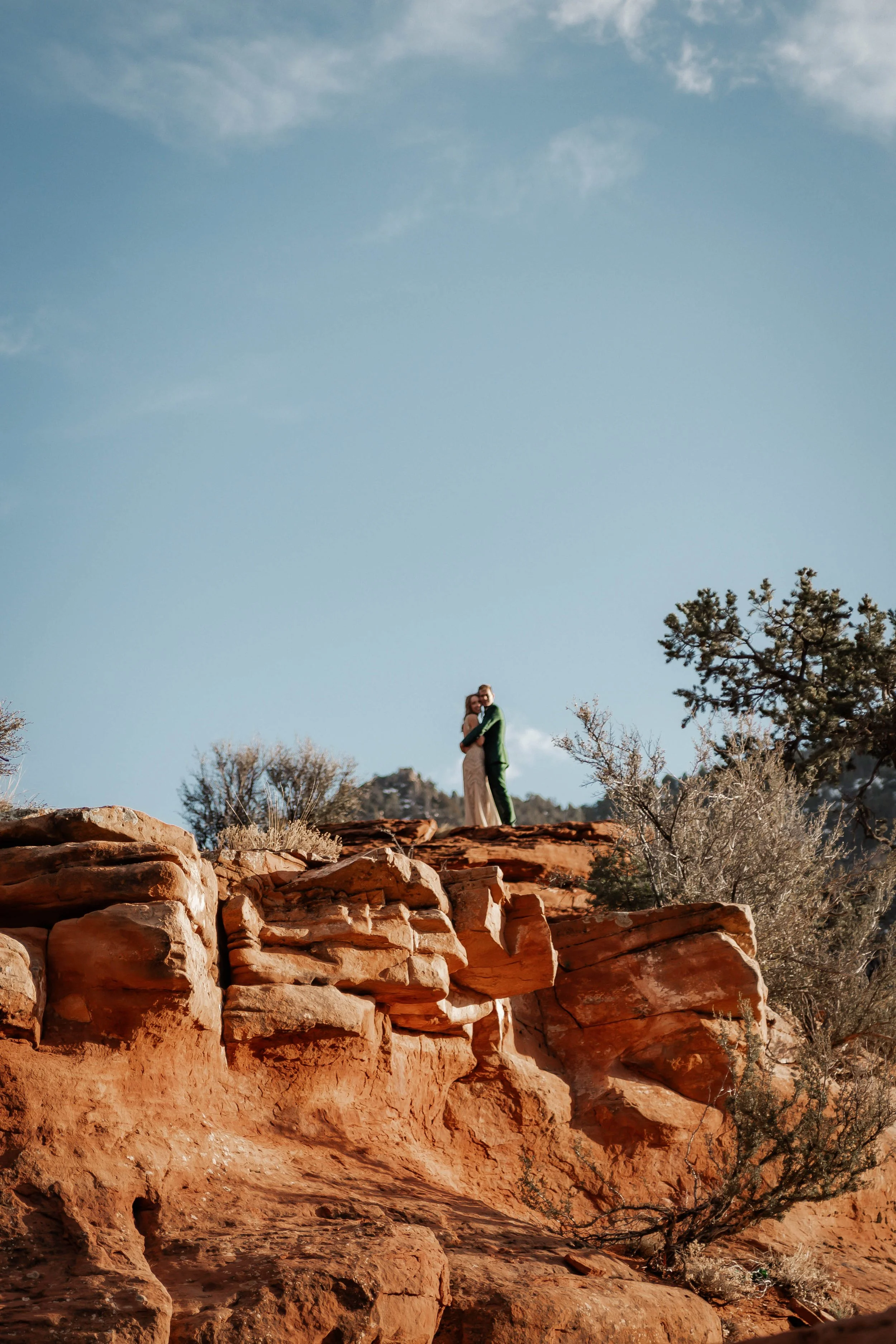 A couple in wedding attire hugging on a rocky desert in Sedona landscape with a clear blue sky.