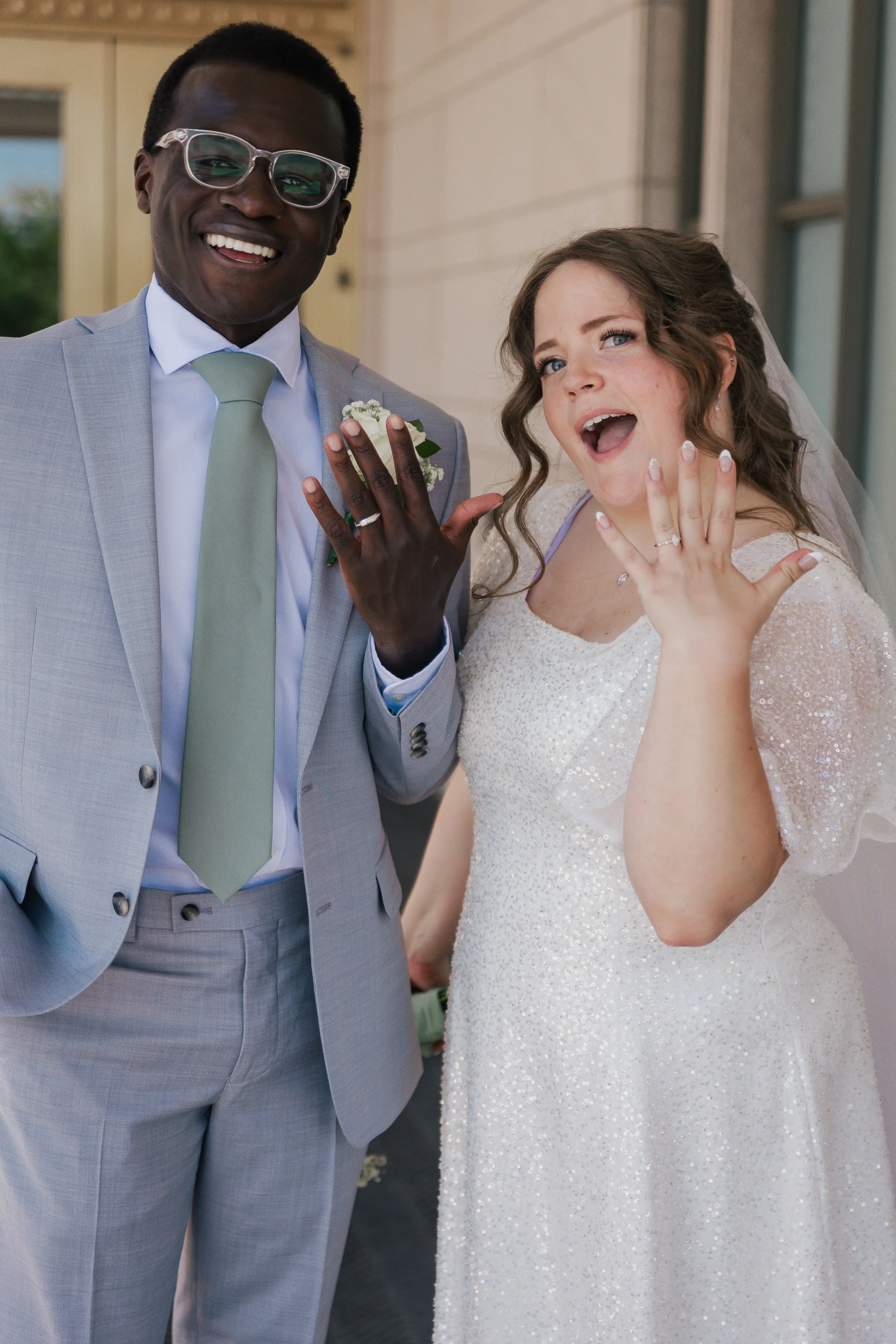 A bride and groom outdoors, showing off their wedding rings, smiling and making a playful gesture.