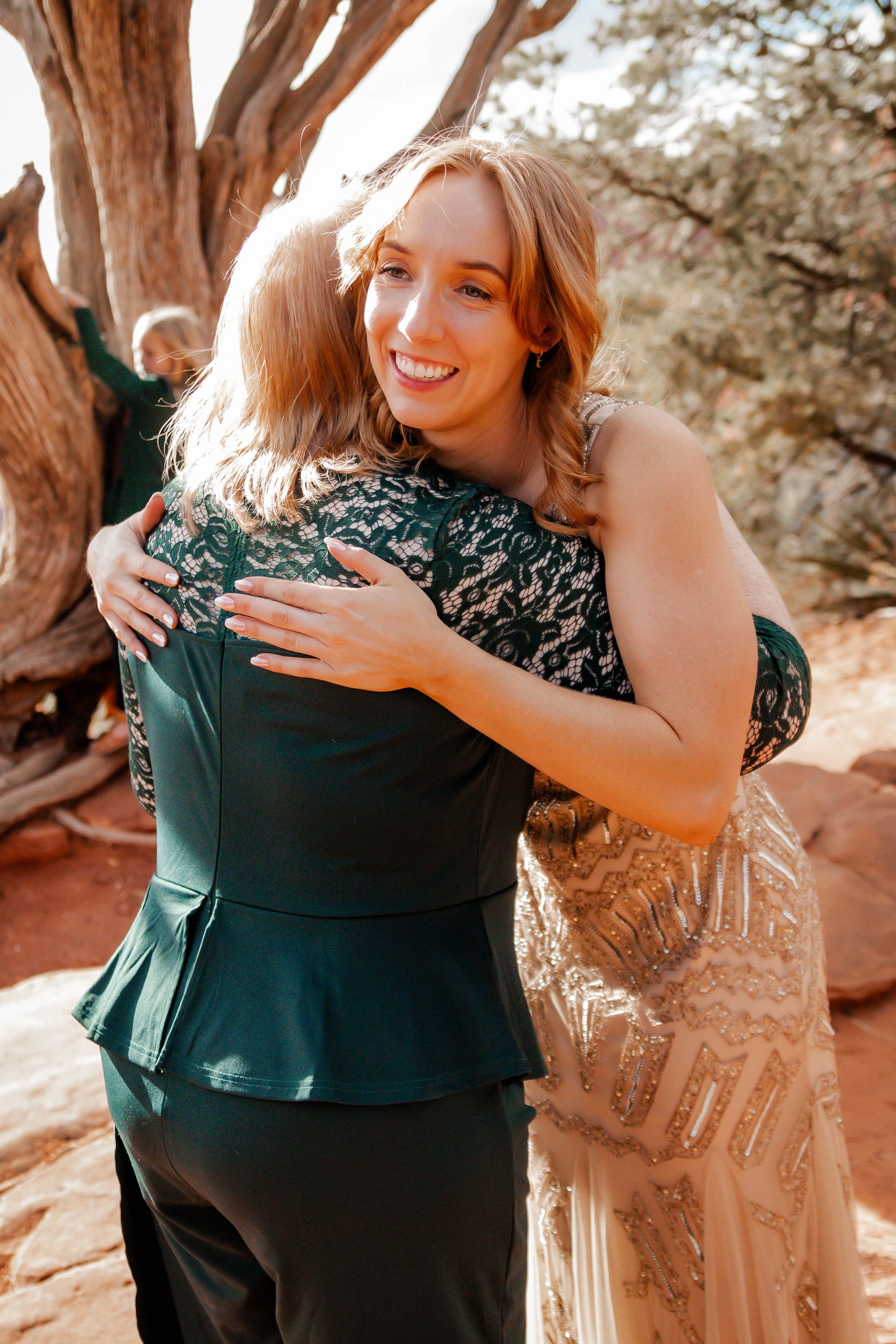 Two women hugging outdoors, one wearing a black lace top and the other in a beige dress with intricate beadwork, near a tree with twisted branches in a natural setting.
