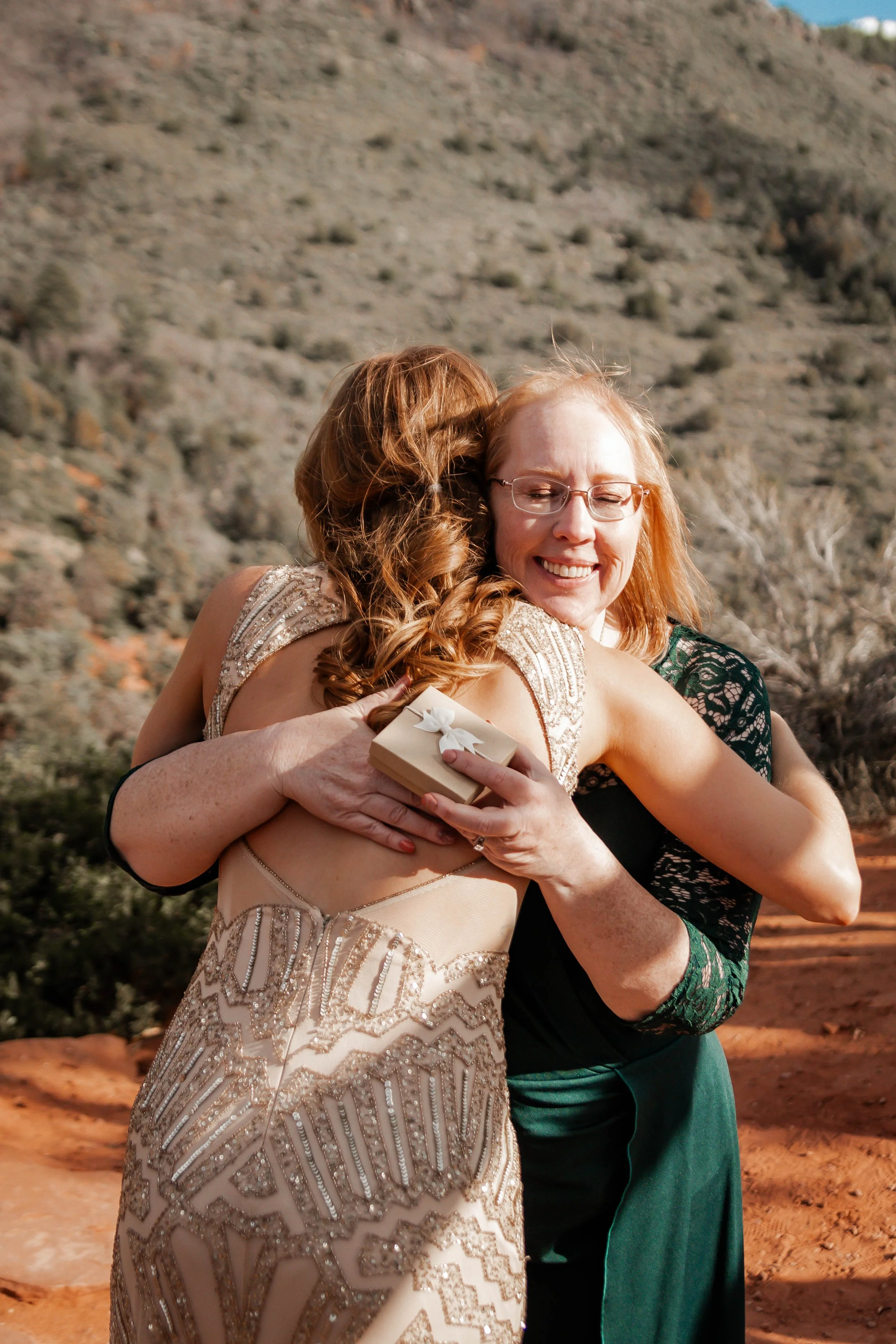 Two women hugging outdoors; one in a formal dress holding a small gift, smiling happily.